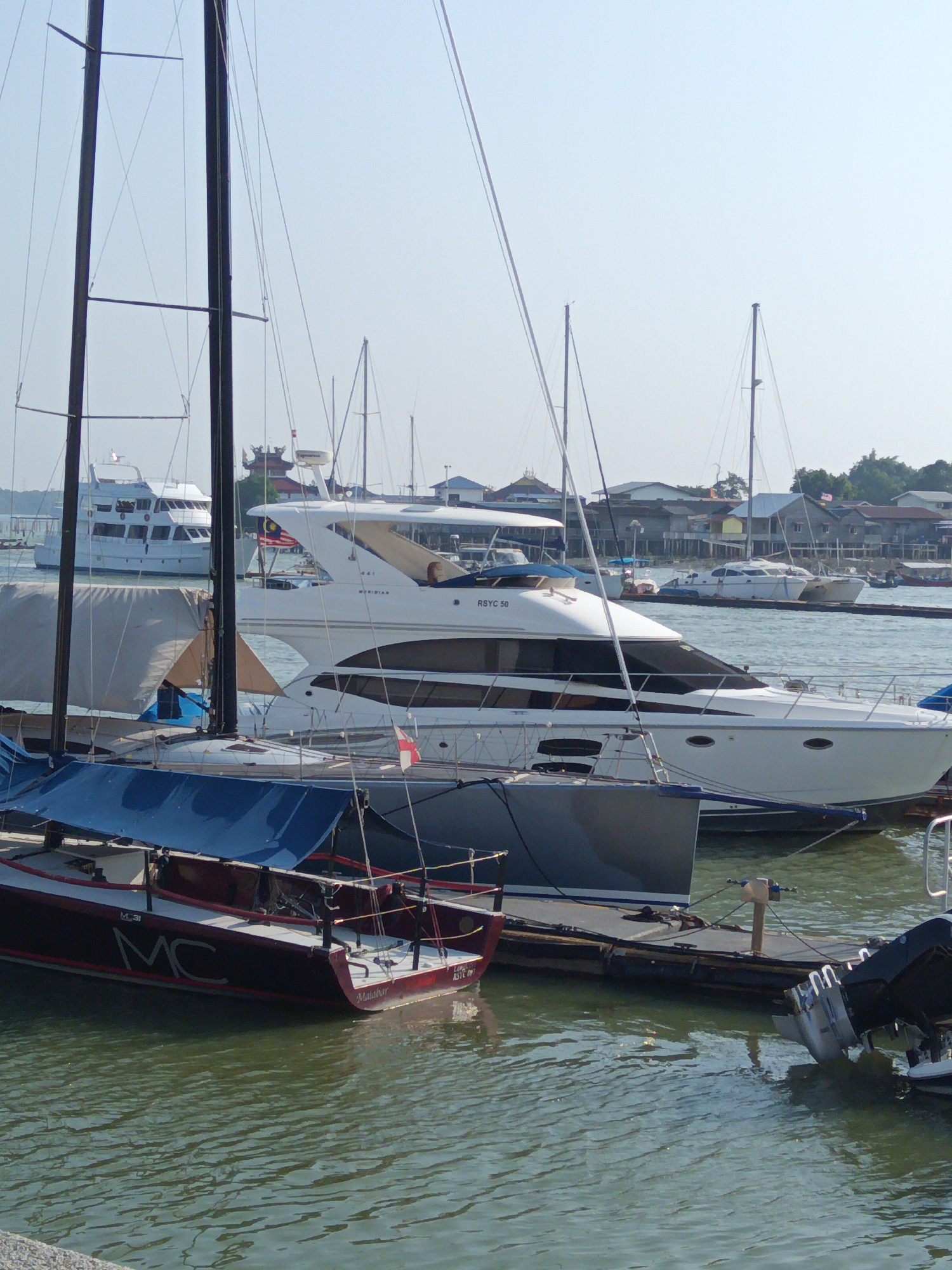 The image shows a marina in Klang City, Malaysia, on a bright, clear day. The foreground features a dark red sailboat with white lettering that reads "MC". Behind it, a larger white yacht with black trim is docked. The yacht has the name "MERIDIAN" visible on its side, along with "RSYC 50". Further back, another large white yacht is visible, and beyond that, a row of buildings with distinct architectural features, possibly traditional wooden houses on stilts, line the shore. Several other sailboats and yachts are moored in the water, their masts creating a complex network against the pale blue sky. The water is calm and reflective, showing the ripples from the boats. The overall scene depicts a busy harbor with various vessels and the characteristic waterfront architecture of Klang City.