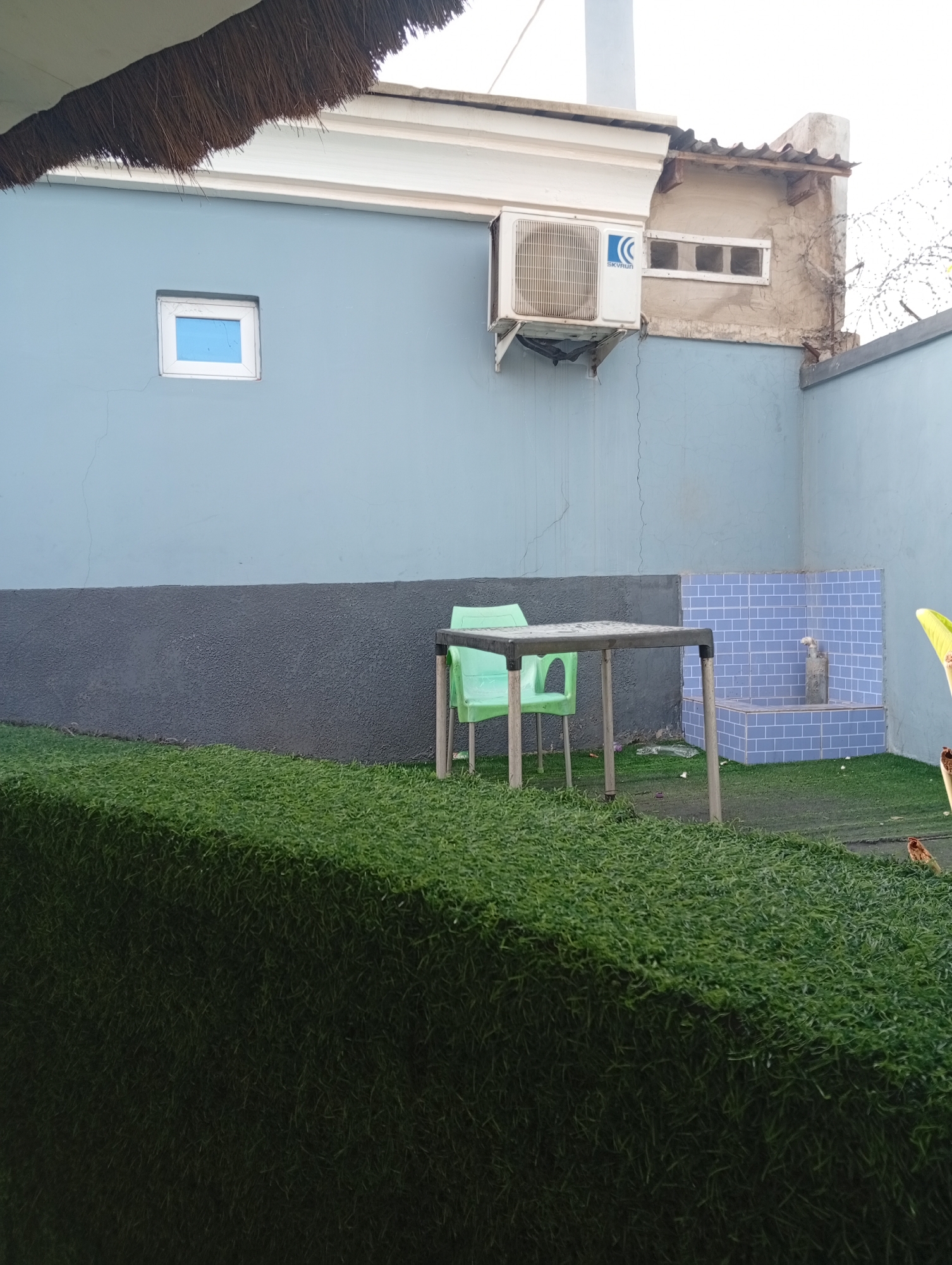 The scene depicts an outdoor space in what appears to be Adankolo, Nigeria. A light blue building forms the backdrop, with a small square window and an air conditioning unit mounted on the exterior wall. A gray textured surface runs along the lower portion of the building. A light green plastic chair sits at a gray plastic table. To the right of the table is a small, tiled water fountain feature. A dense patch of green artificial grass dominates the foreground, obscuring much of the ground level. A bird stands on the edge of the artificial grass, facing right. The scene appears to be a courtyard or small garden space.