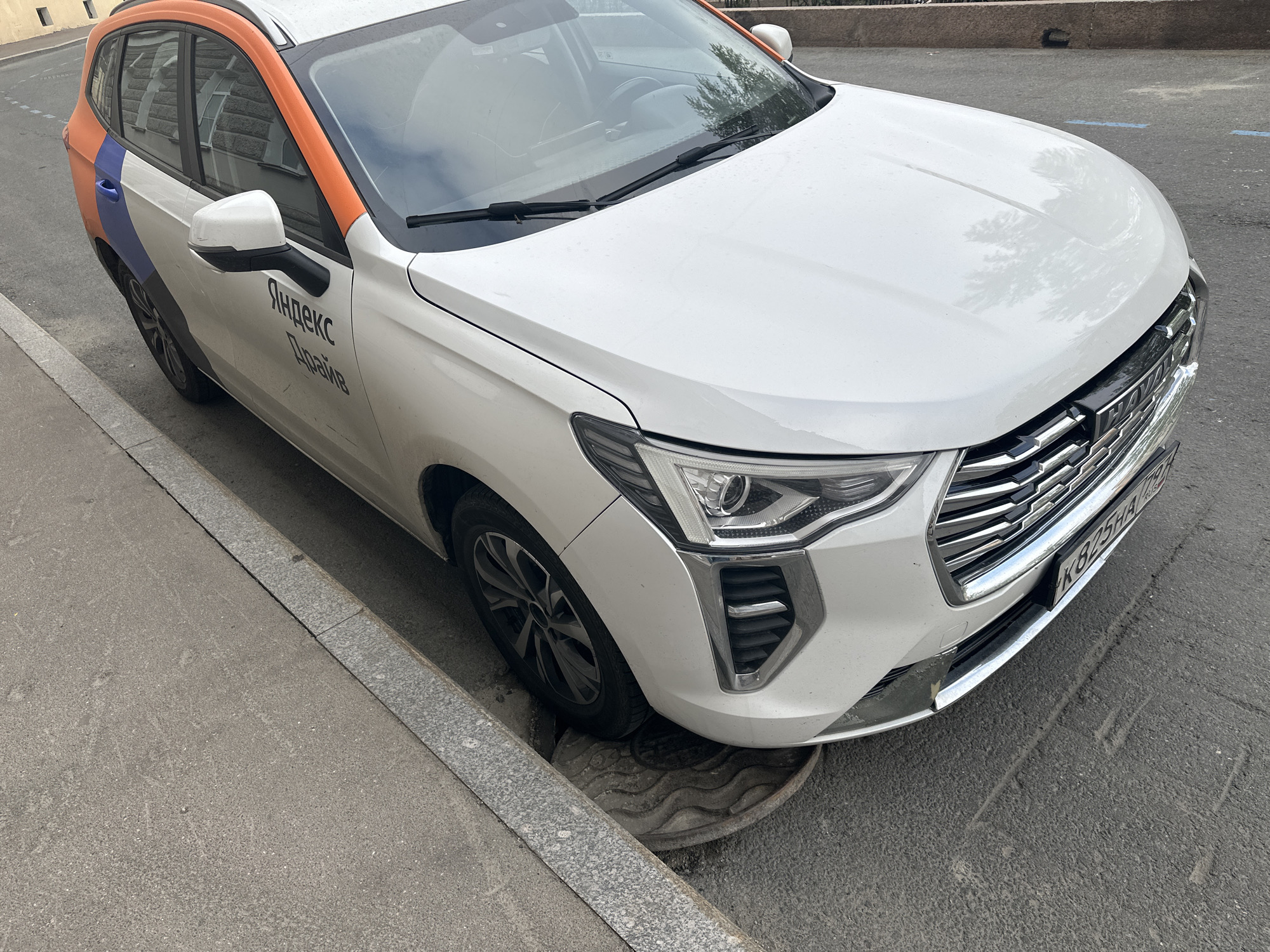 A white Haval car with orange and blue taxi markings is parked on a street in Saint Petersburg, Russia. The car's front right tire is on a manhole cover. The car has "Яндекс Драйв" (Yandex Drive) lettering on the side. The license plate reads "К 825HA 799". The car is parked next to a gray curb. The road surface is asphalt.