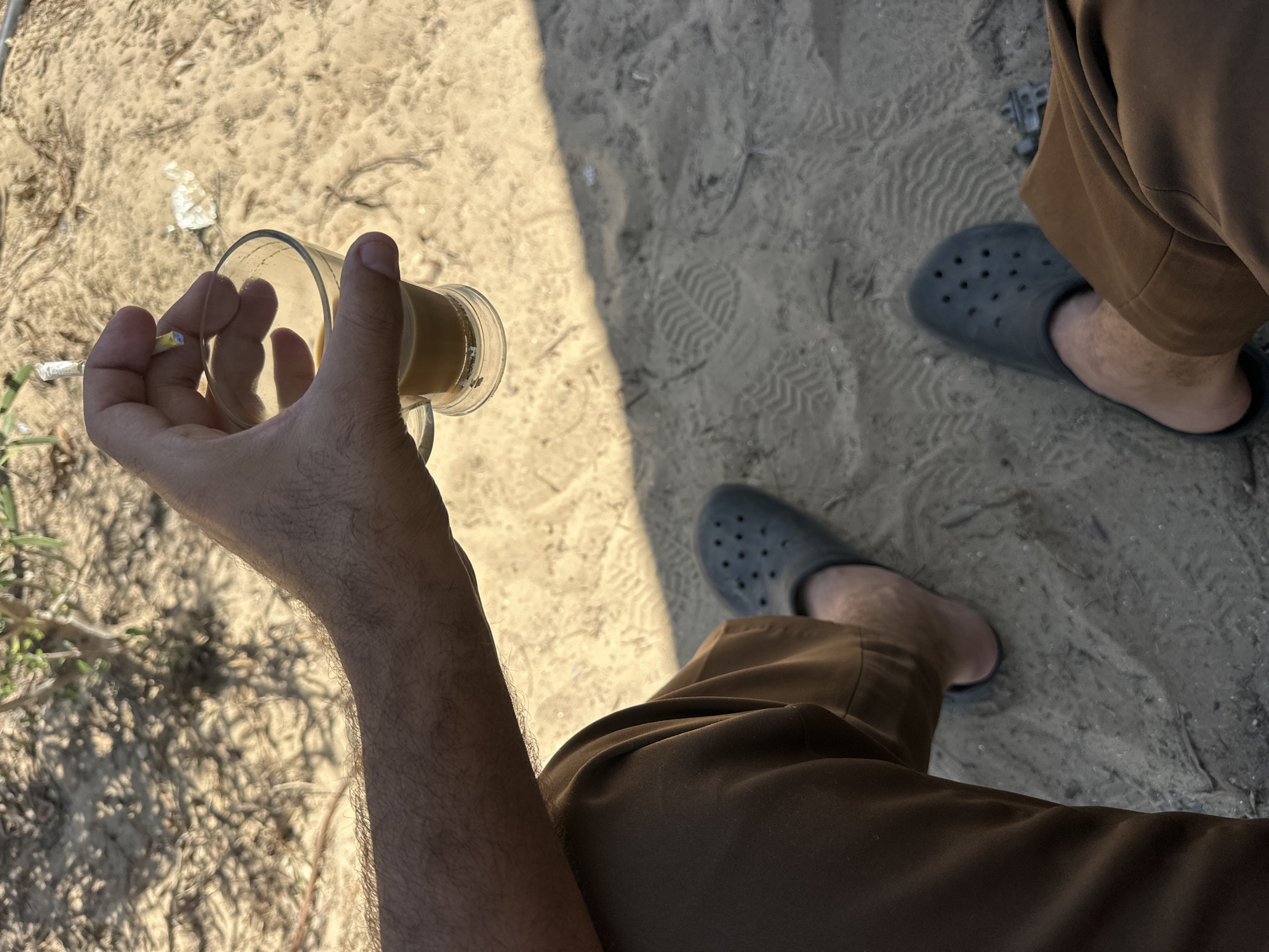 A person's hand holds a glass of tea and a cigarette over a sandy surface. The person is wearing brown pants and gray crocs. The background consists of sand and vegetation. The lighting suggests an outdoor setting during daylight hours.