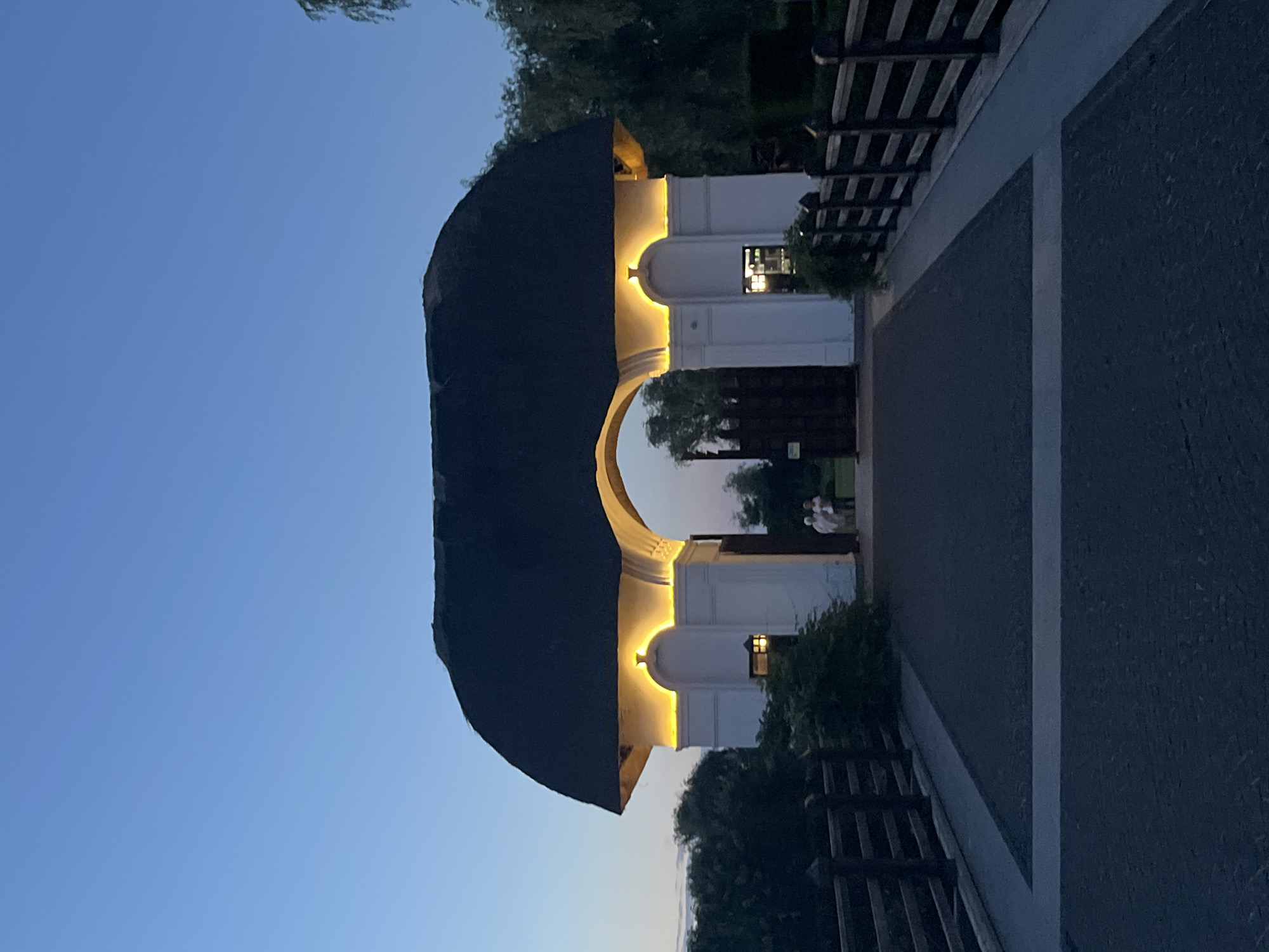A low-angle, wide shot captures an arched gateway structure with a thatched roof against a twilight sky. The gateway is illuminated with warm yellow lights under the eaves. White pillars support the arch, with decorative elements and a small lantern visible on the left pillar. A dark wooden gate stands open within the archway. Two individuals, likely tourists, are standing near the gate. A paved walkway leads to the gateway, flanked by wooden fences on either side. Lush green trees and foliage surround the area, with the sky a clear, pale blue. The scene appears to be an entrance to a park or estate.