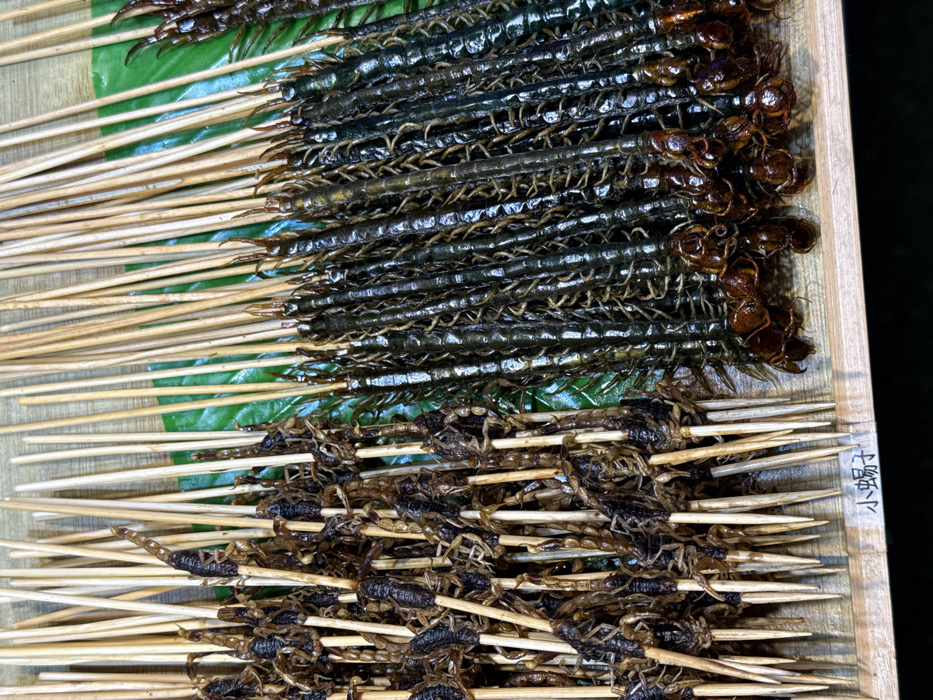 A forensic close-up image depicts a market stall display of skewered arachnids and arthropods, reportedly located in Yuzhong District, China. The left portion of the frame shows approximately fifty small, dark brown scorpions, each impaled lengthwise on a thin wooden skewer. The scorpions exhibit a fried or roasted appearance, with curled tails and visible legs. The right portion features approximately forty long, dark green to black centipedes, similarly impaled on wooden skewers. Their segmented bodies and numerous appendages are distinct, with some centipede heads and tails displaying a reddish-brown hue. All skewers are arranged vertically or leaning, presented on a light-toned wooden surface. A bright green leaf, possibly a banana leaf, partially underlies the centipede skewers. Chinese characters translating to 'small scorpion' are inscribed on the wooden surface in the lower-left quadrant. No human figures are present.