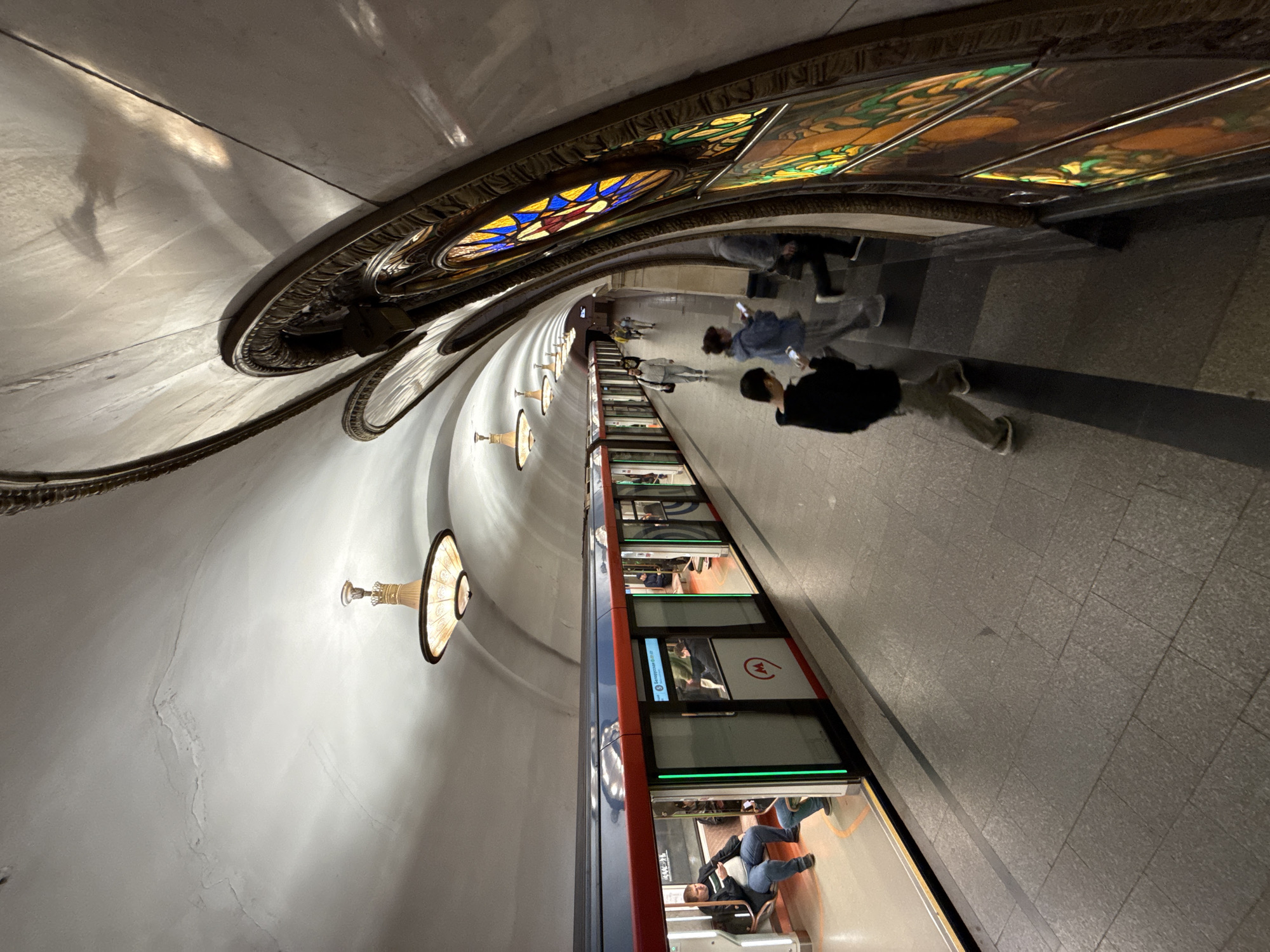 The image depicts the interior of a Moscow Metro station, likely adorned in a grand, ornate style. In the foreground, a boy in a black hoodie and light-colored pants walks away from the camera. To his right, a woman in a blue hoodie and grey pants is visible, also looking towards the right. Further down the platform, several other individuals are walking or waiting for a train.
The station features a vaulted ceiling with decorative light fixtures. Along the right wall, there are large, arched stained-glass windows with colorful geometric patterns, characteristic of some of Moscow's more elaborate metro stations. The platform itself is paved with grey tiles.
On the left side of the image, a train with its doors open is stopped at the platform. The train cars are predominantly dark blue with a red stripe near the top and green accents around the doors. Passengers are visible inside some of the train cars, and one man is seated on a bench near the open door. The train displays the Moscow Metro logo, a red circle with a white "M" inside. The overall ambiance suggests a busy public transportation hub.