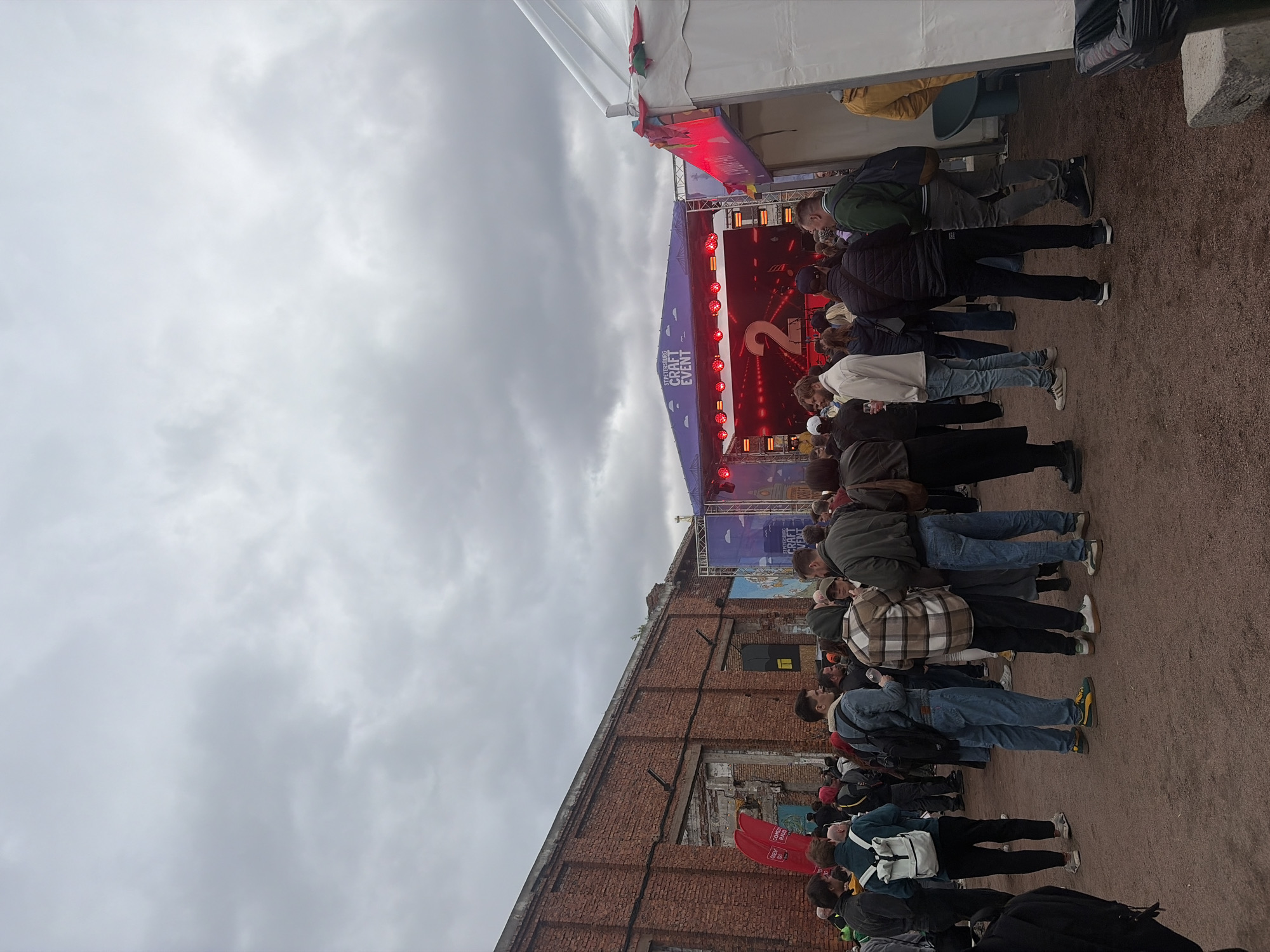 A medium shot shows a crowd of people at the Saint Petersburg Craft Event in Russia. The crowd is looking towards a stage with a red screen with a large number "2" on it. There are tents and an old brick building in the background. The sky is overcast.