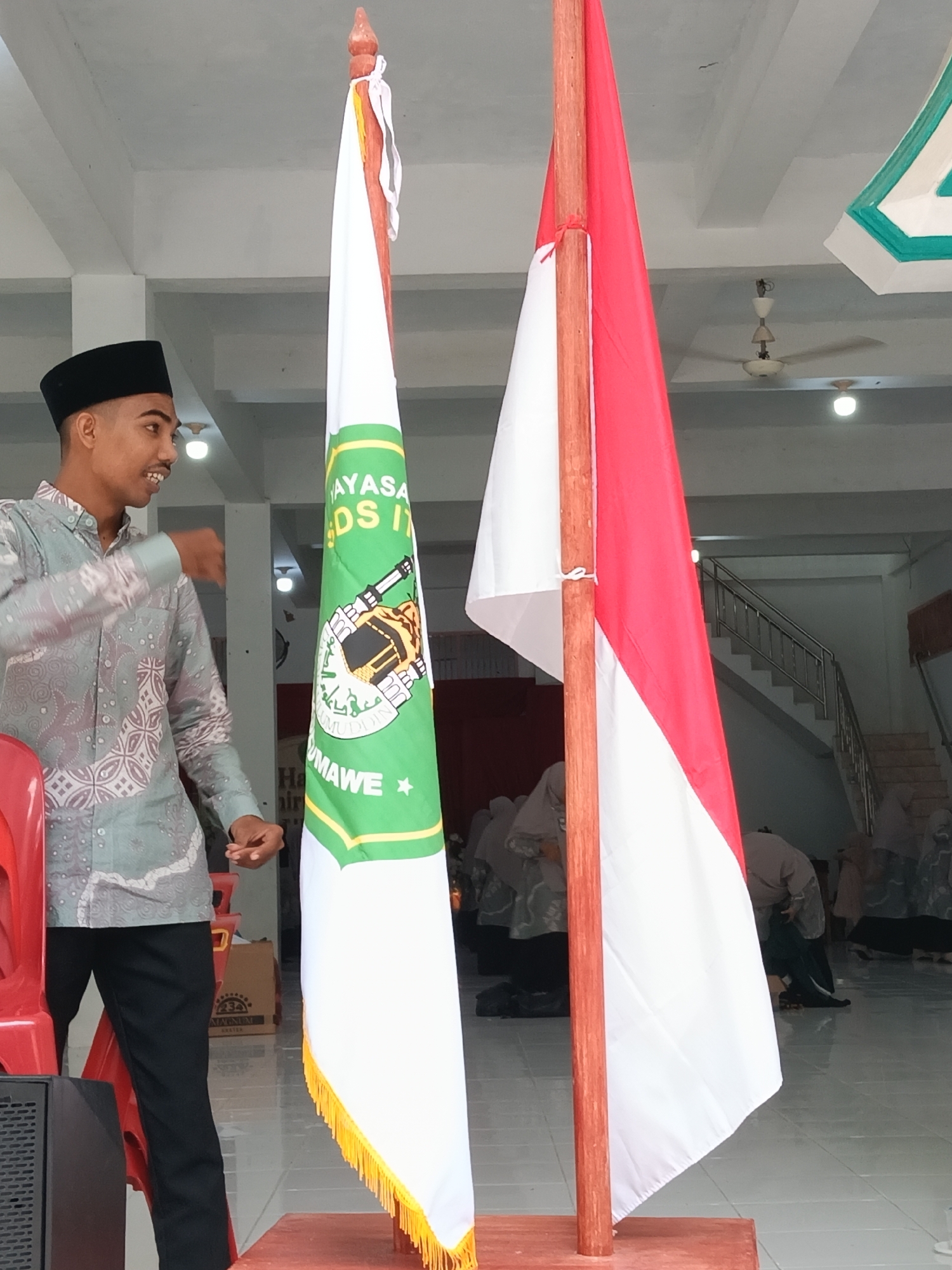 The image depicts a man wearing a patterned grey shirt and black pants, standing next to two flags in what appears to be an indoor space. He is wearing a black peci hat and gesturing with his hands. The flags are mounted on wooden poles and are positioned side by side. The flag on the left is mostly white with a green section featuring the image of the Kaaba. The flag on the right is red and white, resembling the Indonesian flag. The location appears to be in Lhokseumawe, Indonesia. Several people are kneeling on the floor in the background, presumably in prayer. The interior space has white walls, a white tiled floor, and a visible staircase leading to an upper level. There are also visible ceiling fans and light fixtures. A red chair is placed in the foreground on the left side of the image.