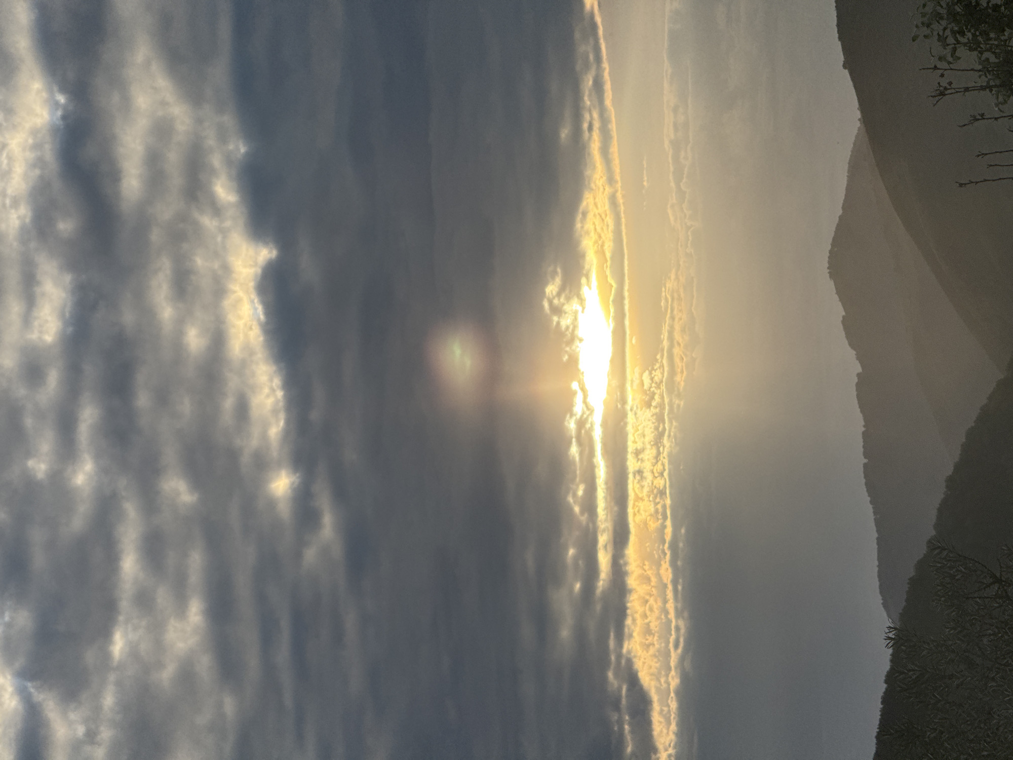 This is a photograph of a dramatic sunset over a mountainous landscape. The sun is low in the sky, casting a bright, golden light that breaks through dark, textured clouds. The light illuminates the edges of the clouds, creating a stark contrast with the shadowy masses. In the foreground, the silhouette of a dark, hazy mountain range rises from the right side of the frame. The mountain's slope is gentle, and its contours are softened by the atmospheric haze. On the far right, a few dark branches of a tree are visible against the lighter sky, adding a touch of foreground detail. The overall impression is one of a powerful natural phenomenon, with the dramatic play of light and shadow evoking a sense of awe and natural beauty. The time of day is clearly sunset, given the low angle of the sun and the warm, golden light. The weather appears to be partly cloudy, with the sun managing to break through. The location is identified as Soloneshnoye, Russia.