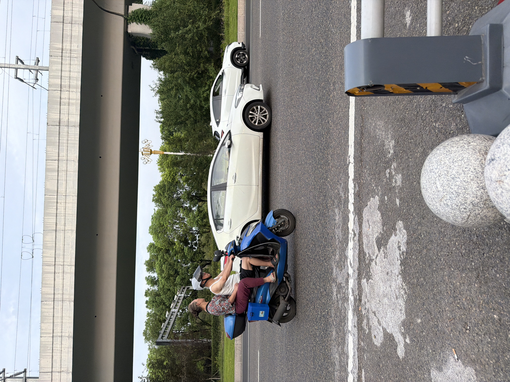 A man and a woman are riding on a blue scooter on a road in Jiangjin, China. The man is driving and wearing a white tank top and black shorts, and a helmet. The woman is sitting behind him and wearing a patterned top and maroon pants. The scooter has a blue storage box on the back. In the background, there are trees, a white car, and an elevated highway. The sky is cloudy. The foreground shows the asphalt road with white lines and some white patches. There are also some large, speckled spheres and a gray traffic barrier with yellow and black stripes.