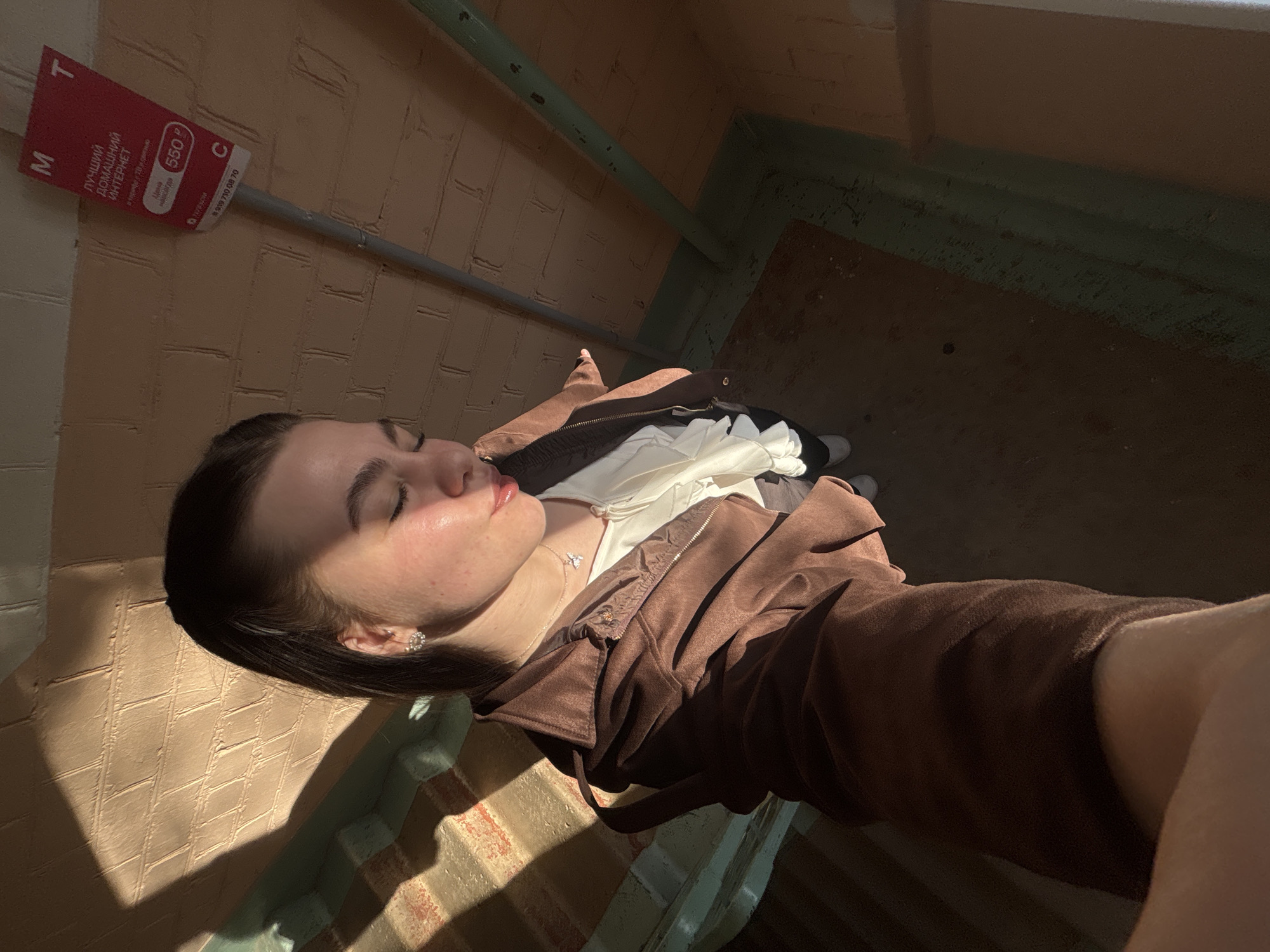 A selfie photograph shows a young woman with fair skin and dark hair lying down, possibly on a stairway or landing. She is wearing a brown zip-up jacket over a white blouse, with visible jewelry including earrings and a necklace. The background suggests an interior space, with light peach brick walls and a green-painted handrail. The location can be identified as Perm, Russia, based on a red advertisement sign that reads "Лучший Домашний Интернет" (Best Home Internet) in Russian, which is a service offered by the Russian telecom company MTS. The photo is taken at an angle, creating a perspective where the subject's face and upper body are close to the camera. Sunlight creates strong shadows on the wall, adding to the contrast in the image.