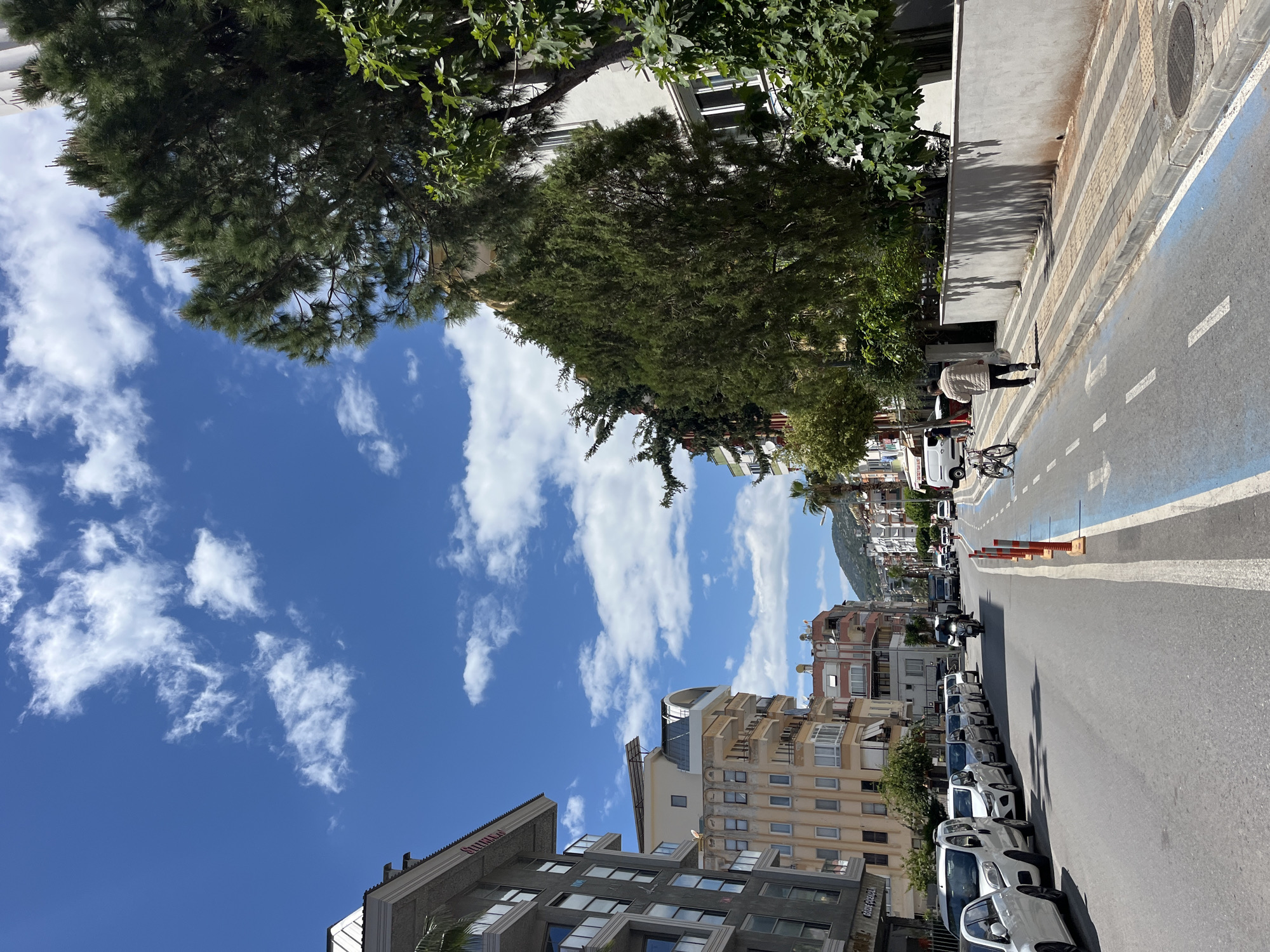The image depicts a street scene in Alanya, Turkey, under a bright blue sky with scattered white clouds. The street runs horizontally, with buildings lining the right side of the frame. Several cars are parked along the roadside, and a motorcycle is seen further down the street. On the left, there's a sidewalk with a blue bike lane. A person in a light-colored jacket is walking along the sidewalk, and a bicycle is parked nearby. Trees and foliage are present, casting shadows on the sidewalk and buildings. In the distance, a hill or mountain is visible. The overall impression is of a sunny day in a moderately urban environment.
