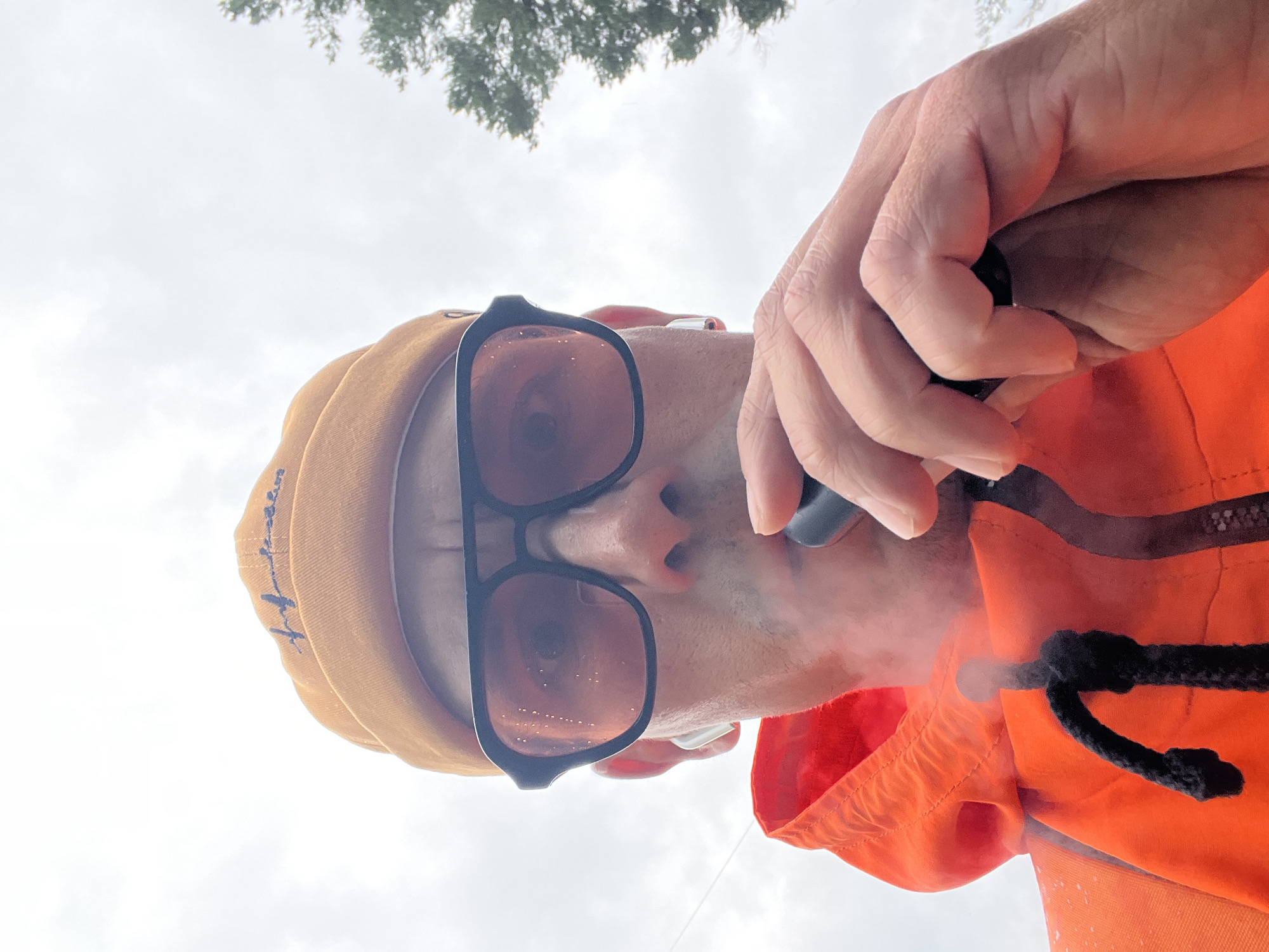 A man is captured in a low-angle, close-up shot, wearing an orange vest over a tan beanie and dark-rimmed sunglasses. He is holding a black object, possibly an e-cigarette, to his mouth, and a plume of vapor emanates from it, obscuring part of his face. His right hand is visible, holding the object. The background consists of an overcast sky and the tops of green trees, suggesting an outdoor setting. The image has an orange cast, likely due to the lighting or reflections from his vest. The man's expression is unreadable behind the sunglasses and vapor. The composition is dominated by the man's face and upper body, with the sky and foliage providing a muted backdrop. No text is clearly visible.