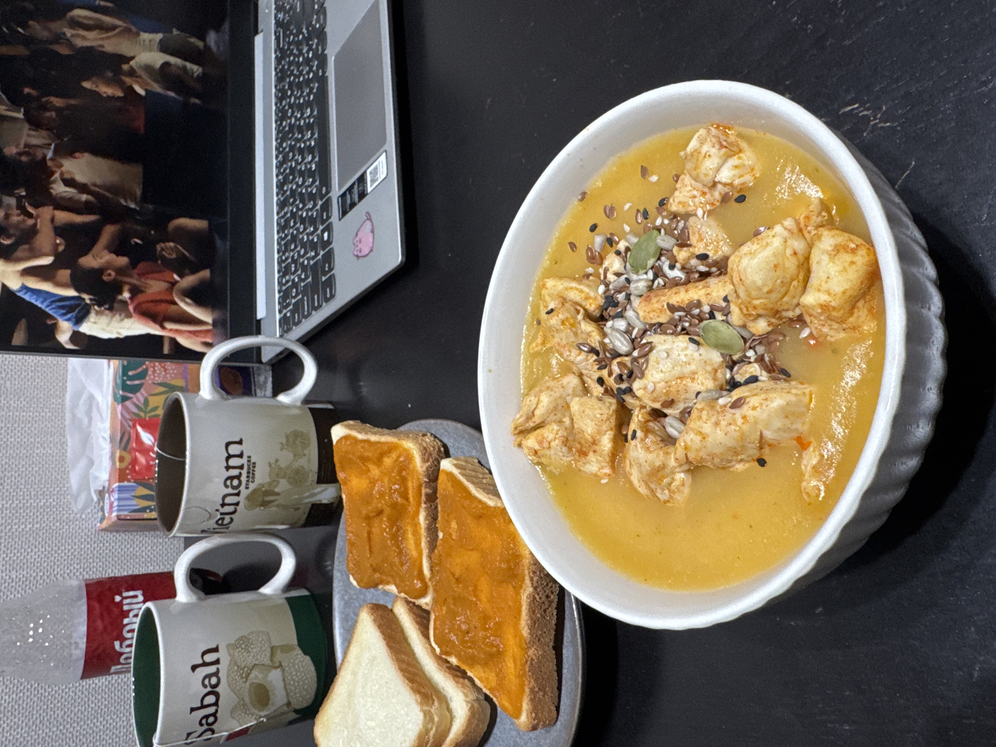 A table with various items is seen. The table surface appears to be a dark grey color. On the table are a laptop, two mugs, a plate with bread, and a bowl of soup with pieces of chicken and seeds on top. One mug has the word "Vietnam" on it. The other mug says "Sabah." A clear bottle and a red box, labeled in Russian, are partially visible behind the mugs. On the laptop screen, a crowd of people can be seen. The location is Perm, Russia.