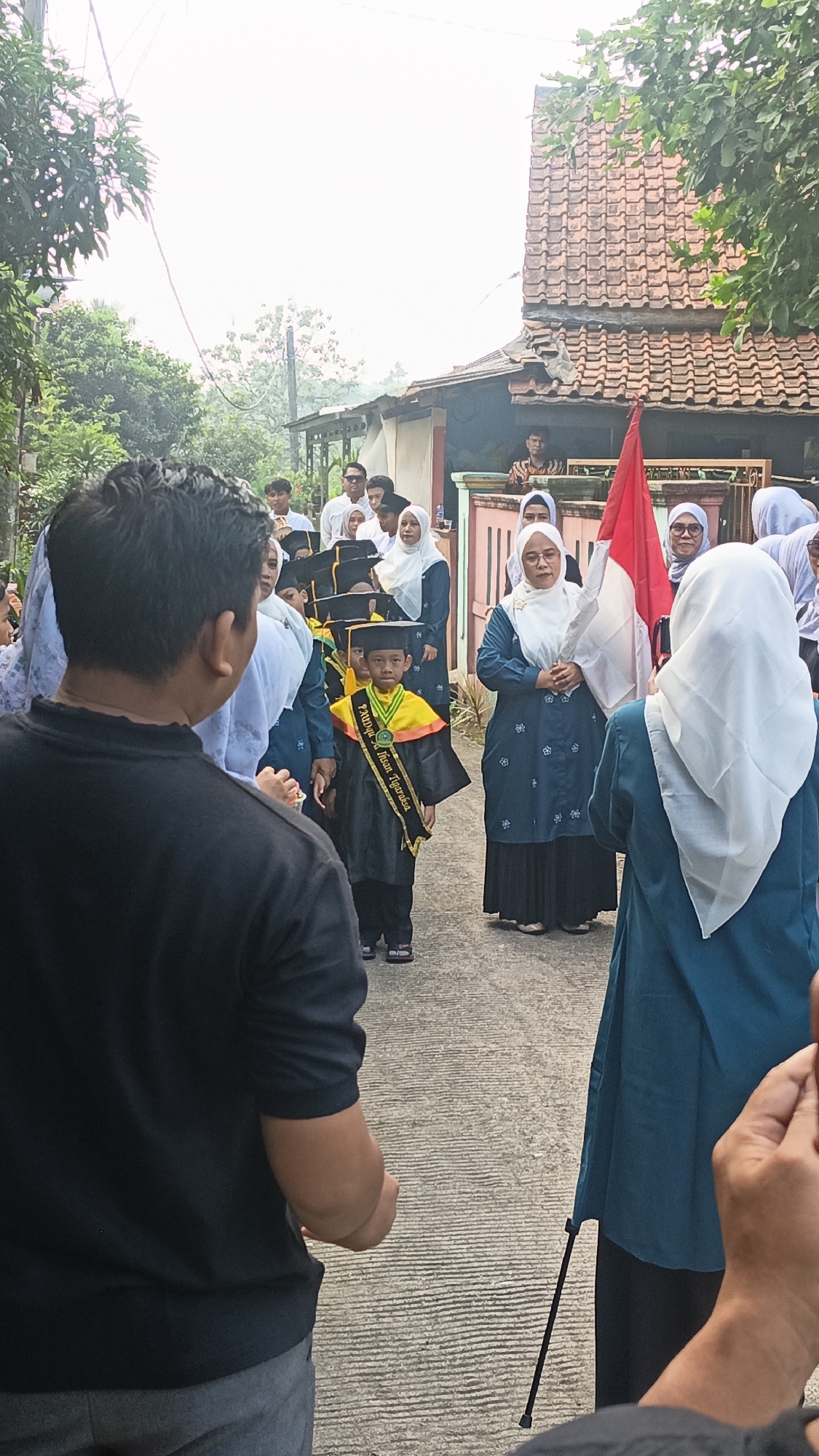 In Tigaraksa, Indonesia, a graduation procession is underway in a residential area. A young boy, wearing a graduation cap and gown with a sash that reads "PAUD Harapan Tiga Raksa," walks down a paved path lined with houses. He is accompanied by adults, some of whom are also wearing traditional Muslim attire, including hijabs. A woman in the foreground holds a red and white Indonesian flag, suggesting a patriotic or celebratory event. The environment appears to be a suburban street with trees and modest houses visible in the background. The scene suggests a community event celebrating the achievements of the children.