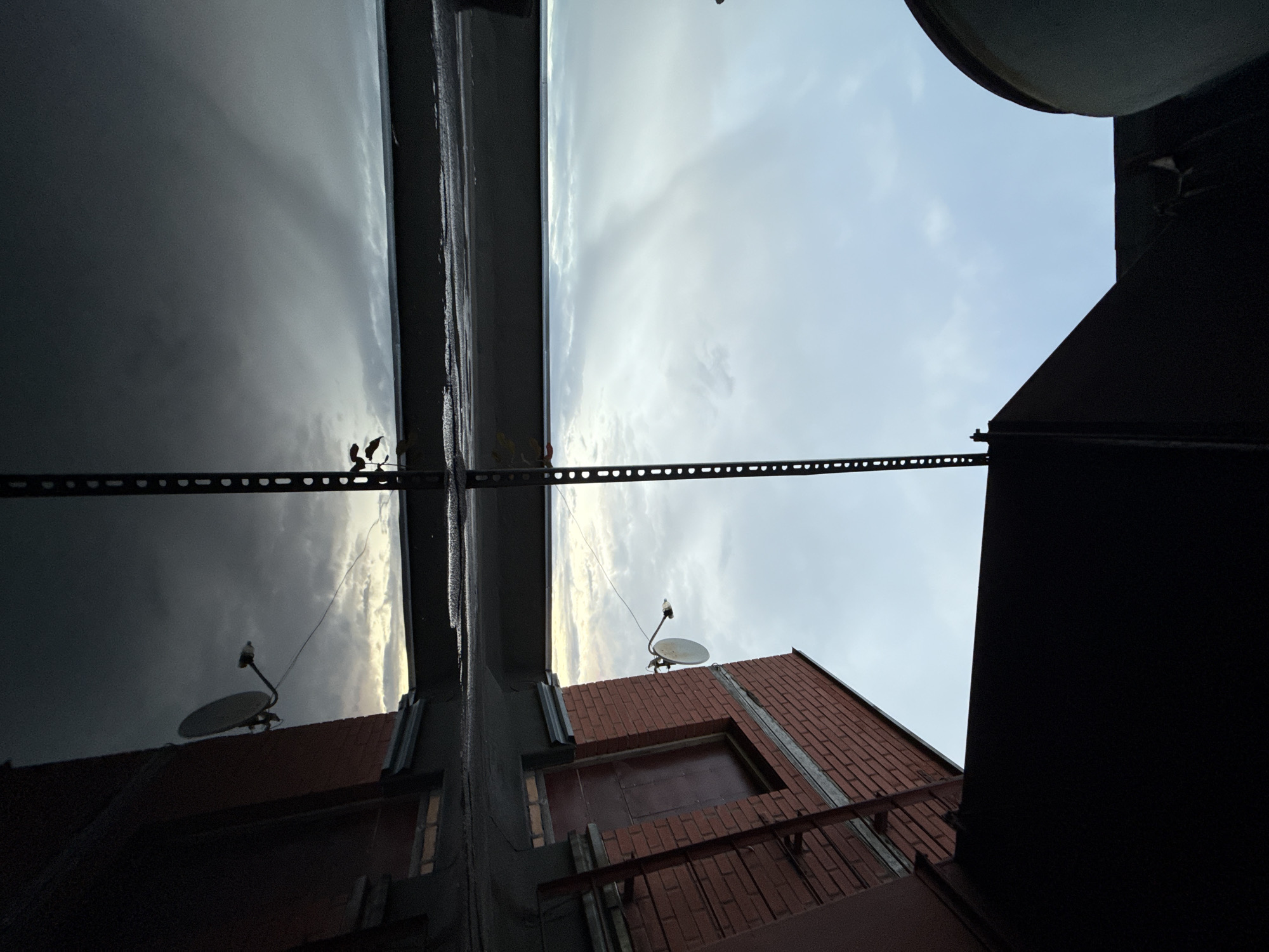 The media file captures an unusual perspective of an urban rooftop scene in Khimki, Russia, likely taken during a cloudy or overcast day. The camera is positioned looking upwards, giving a disorienting view that emphasizes the sky and the structures above.
The primary subjects are architectural elements: a dark, angular structure dominates the right side of the frame, partially obscuring the view. To the left, a brick building with multiple windows is visible, topped with what appears to be roofing material. A prominent feature on the brick building's roof is a satellite dish. Across the scene, a metal grate or railing runs horizontally, creating a dividing line.
A single, small bird is perched on the arm of the satellite dish, a solitary figure against the expansive sky. The sky itself is a mix of dark, brooding clouds on the left, possibly indicative of approaching rain or a storm, and lighter, brighter clouds towards the right. The overall mood is somewhat dramatic and perhaps a little melancholic, due to the overcast sky and the stark architectural lines.
There are no visible people or text in the media file. The time of day appears to be late afternoon or early evening, given the subdued lighting and the quality of the sky. The location cues, such as the architecture and the satellite dish, are consistent with a residential or commercial rooftop in an urban environment like Khimki. The reflection of the sky and the structures in a wet surface, possibly the roof itself, adds another layer of visual interest to the scene.