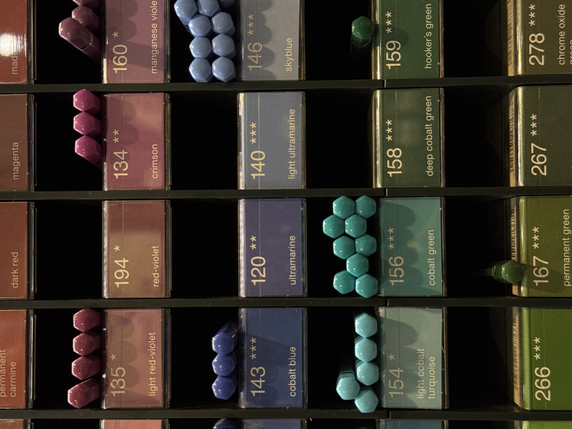 The image displays a grid of art supplies, specifically colored pencils or pastels, organized in vertical columns and horizontal rows. Each item is housed in a labeled compartment, with a number and color name printed on the label. The labels are primarily in English, with some numbers and symbols. The items are various shades of purple, blue, green, and some reddish-brown hues.
The specific location of Moscow, Russia, is not indicated in the image. Therefore, it is not possible to provide details about the city. The scene is an indoor retail or storage environment, likely a stationery store or an artist's supply shop. There are no people or interactions depicted in the image. The focus is solely on the organized display of art materials.