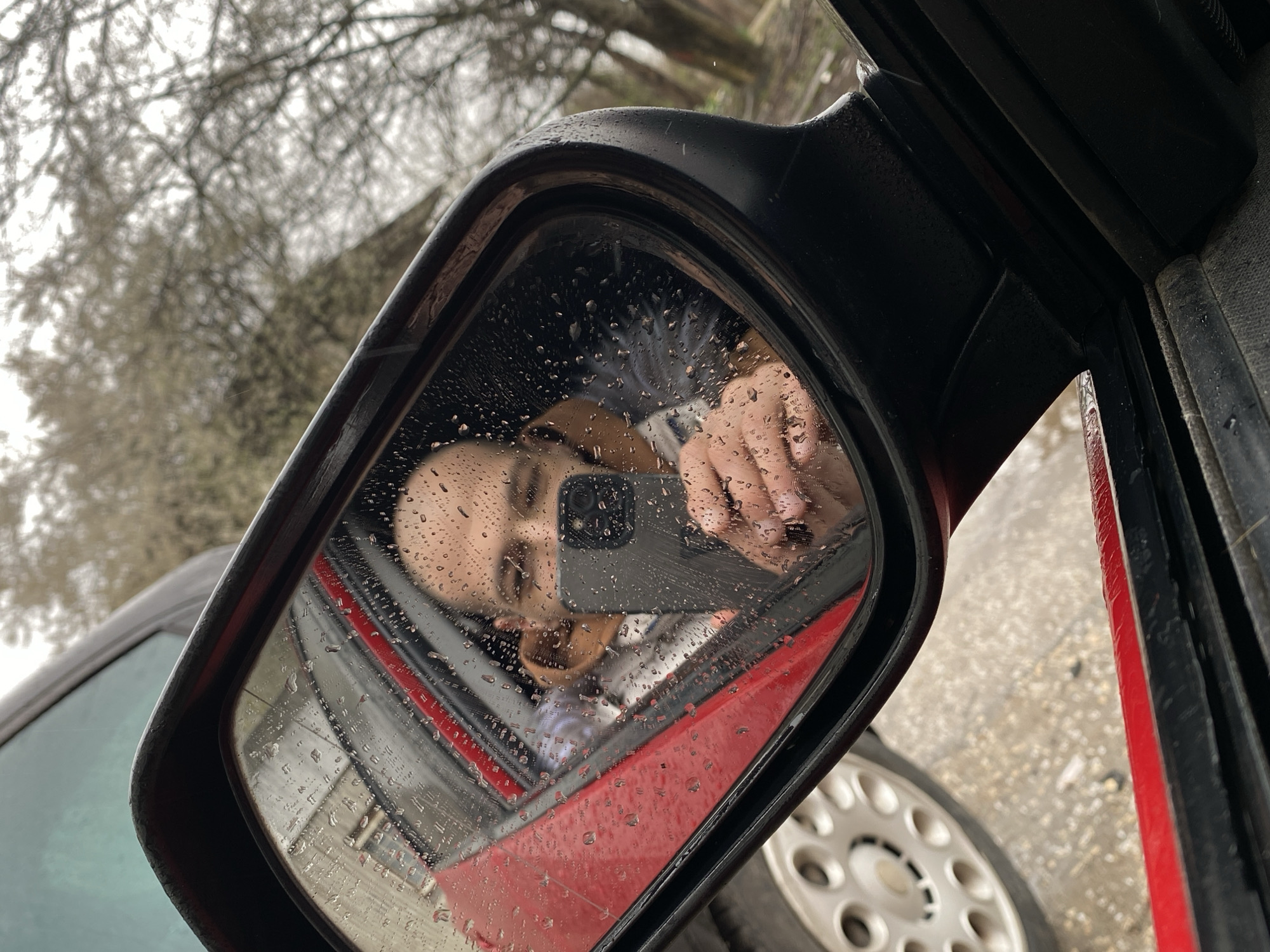 The image depicts a car's side mirror on a rainy day. The reflection in the mirror shows a person holding a smartphone, presumably taking a picture of themselves. Water droplets cover the mirror's surface. The car is red and black. The background includes trees and a wet, paved surface. It is not possible to determine that this image came from Pleven, Bulgaria.