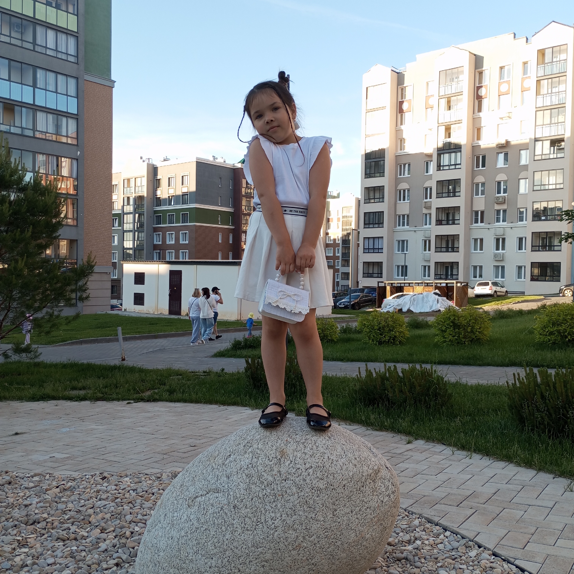 A young girl stands on a large, light-colored, textured boulder in a courtyard with modern apartment buildings in the background. The girl is wearing a white sleeveless top, a white pleated skirt, and black Mary Jane shoes. She is holding a small, white, beaded handbag with a bow on the front. Her hair is pulled up into a bun with two strands hanging down. The apartment buildings are multi-story structures with beige and grey facades, featuring balconies and numerous windows. The courtyard is paved with light-colored interlocking stones and has patches of green grass and manicured bushes. In the middle ground, a small utility building with a dark door is visible, and several adults are walking on the pathway. Further in the background, cars are parked, and a small child in a yellow outfit is walking. The sky is clear and blue, indicating daytime. The scene is set in Pervomaysky, Russia.