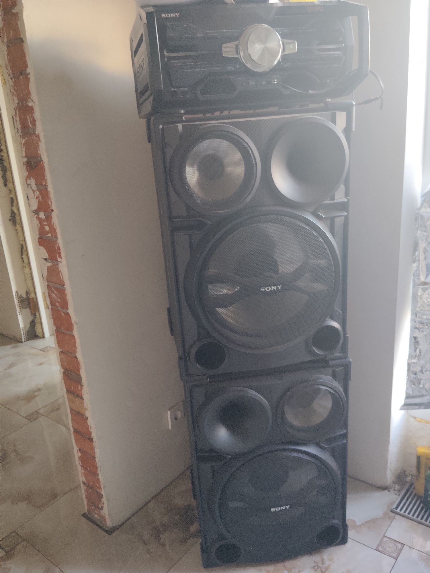 A tall, black Sony stereo system is set against a pale, textured wall. The stereo unit is composed of multiple stacked sections. The topmost section appears to be a CD player and receiver with a prominent silver dial and various buttons. Below this are several speaker units. The largest speaker, positioned in the middle, has a circular speaker grille with the "SONY" logo visible in white lettering. There are other smaller circular speaker drivers and what appear to be bass reflex ports on the other speaker sections. The overall impression is of a powerful audio system.
The scene is indoors, likely a room undergoing renovation. To the left of the stereo, a brick wall is exposed, showing unfinished plaster and insulation. The floor is tiled with a beige and brown patterned design. The lighting suggests it is daytime, with natural light illuminating the right side of the stereo system, creating some glare and bright spots. A white electrical outlet is visible on the wall behind the stereo. To the right of the stereo, some objects on the floor are partially visible, including what might be cleaning supplies or tools, and a striped mat. The setting appears to be a domestic or commercial space in the process of being worked on. No people are visible in the image.