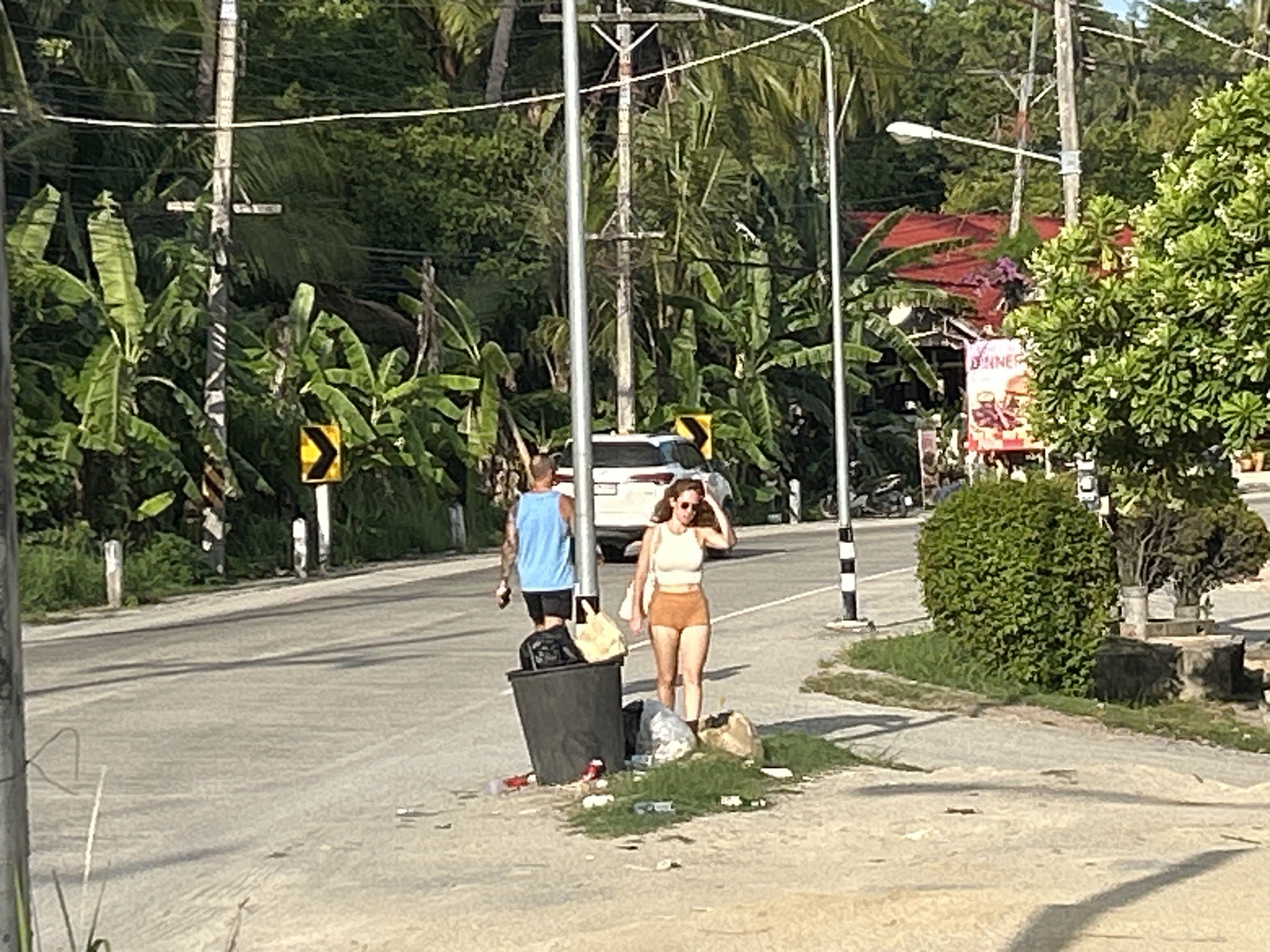 The image depicts a street scene in ตำบลเกาะพะงัน, Thailand. A woman in a light top and orange shorts walks past a black trash bin on a paved area adjacent to a road. To her left, a man in a blue tank top and black shorts walks in the same direction. A white car is parked behind them on the road. The environment consists of lush tropical foliage, including banana trees. Utility poles with overhead wires are visible, and a street light is present. Litter is scattered around the trash bin. A restaurant or establishment with signage is seen in the background.