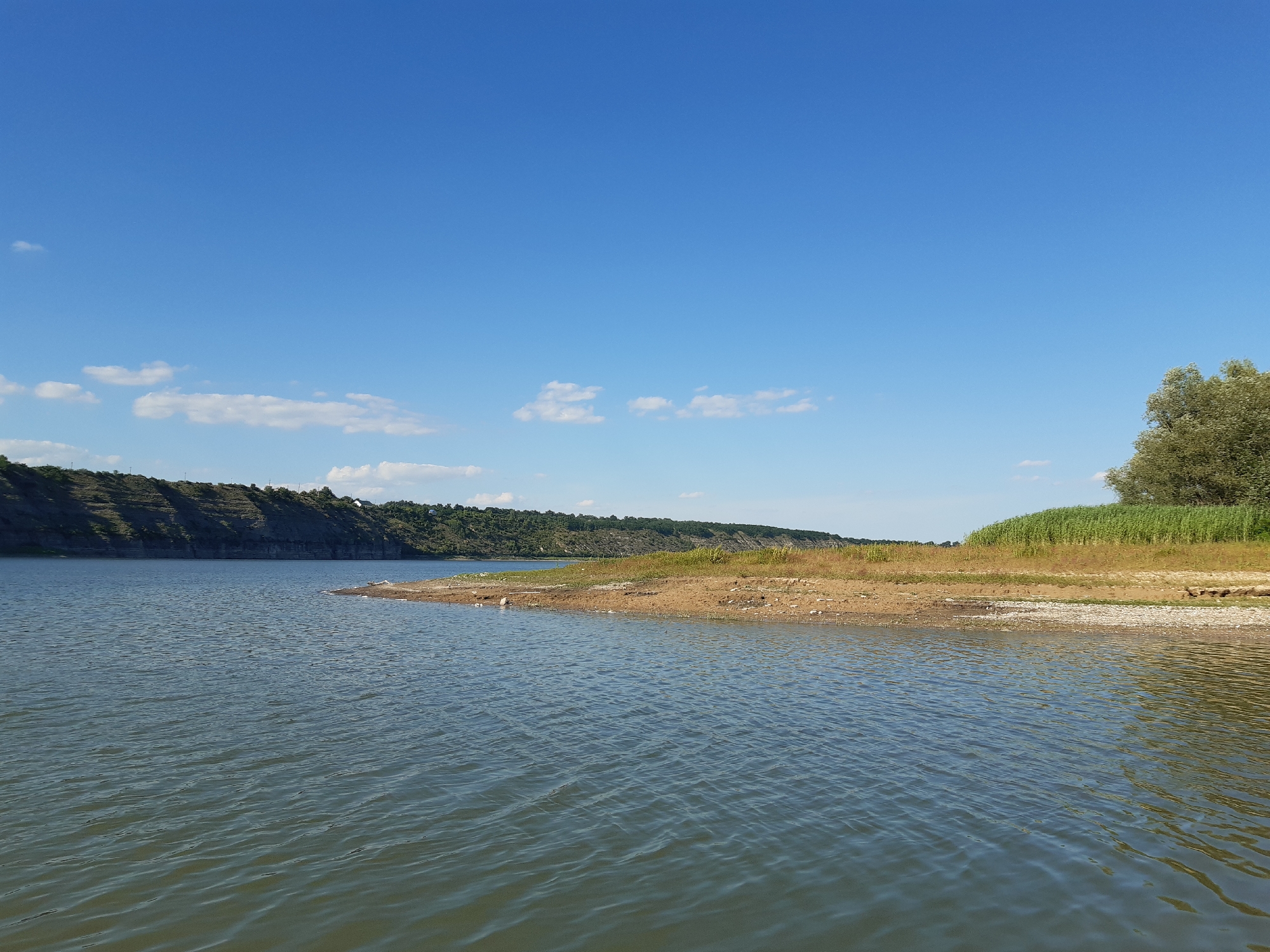 The image shows a wide, calm body of water, likely a river or lake, under a clear blue sky with sparse, wispy clouds. The foreground is dominated by the water's surface, reflecting the sky and showing subtle ripples. To the right, a dry, sandy bank leads to an area of dry grass and reeds. Further back, a lush green tree stands on the bank. The background features a sloping, rocky hillside covered in vegetation, with a few buildings visible on its crest. The overall impression is of a natural landscape on a sunny day. The location is identified as the city of Sokil, Ukraine.