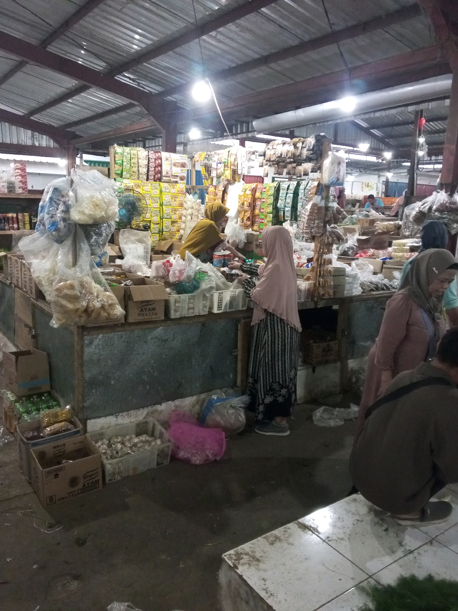 The image depicts a bustling market scene in Gentan, Indonesia. The market is covered by a corrugated metal roof supported by a metal frame, with visible lighting fixtures illuminating the area. In the foreground, several stalls display a variety of goods, including packaged food items, what appear to be snacks or crackers in plastic bags, and boxes of produce.
Multiple individuals are present, engaged in shopping and selling activities. One woman, wearing a yellow hijab and a patterned dress, is standing behind a stall, interacting with customers or arranging merchandise. Another woman, in a light pink hijab and a dark patterned skirt, is seen in profile, also near a stall. A third woman, wearing a gray hijab and a salmon-colored top, is partially visible in the right mid-ground. The lower right portion of the image shows a person in a dark-colored hooded jacket, crouching down.
The ground is concrete, with some areas appearing wet. Several cardboard boxes are stacked, containing various items, including what look like green bottles or cans. A pink plastic bag lies on the ground near some crates filled with small, round objects, possibly garlic or onions. The overall impression is of a busy, local market with an abundance of goods and people interacting within a covered, somewhat cluttered environment.