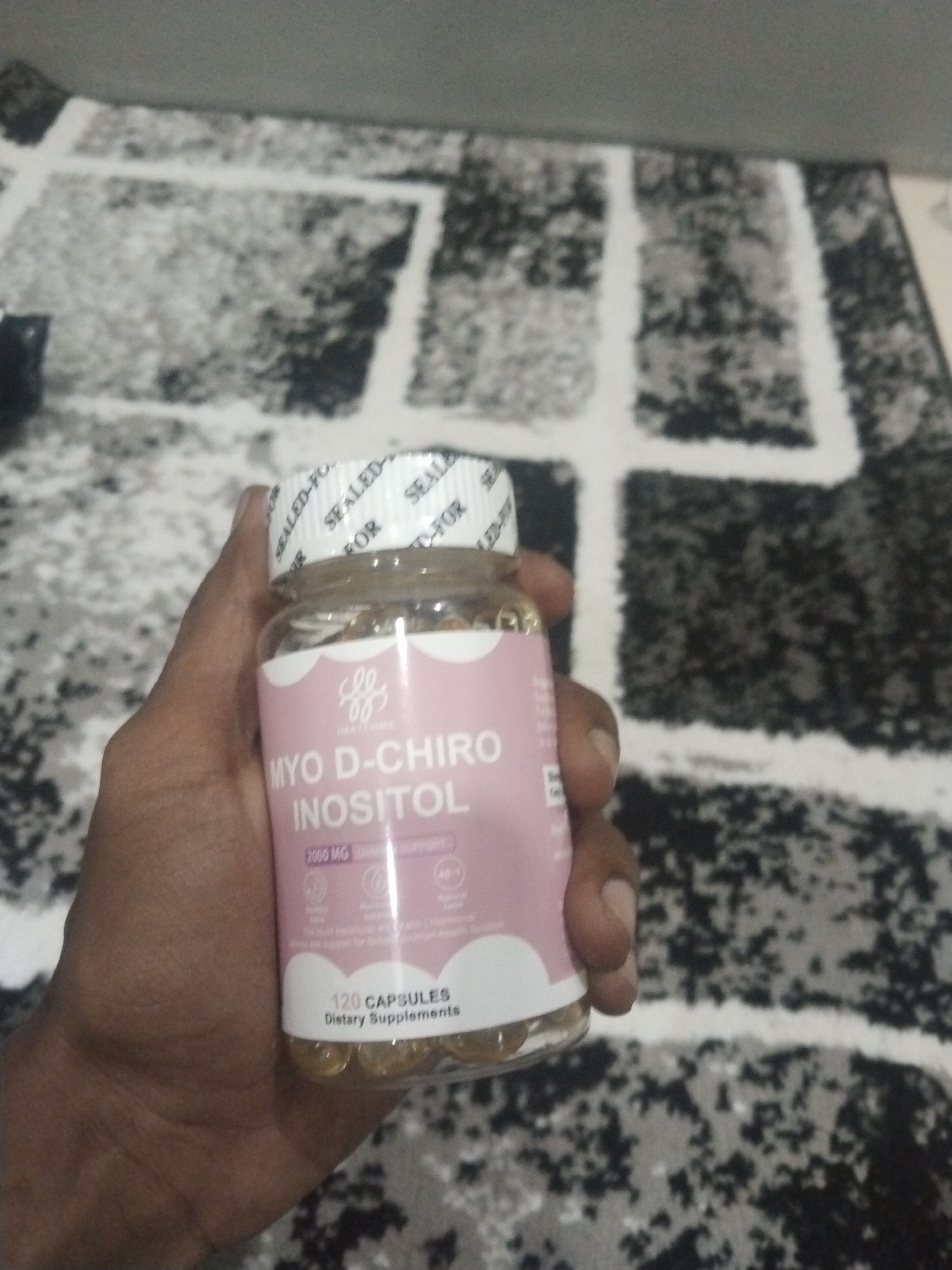 A dark-skinned hand holds a clear plastic bottle of "MYO D-CHIRO INOSITOL" capsules against a background of a patterned rug with white, gray, and black sections. The bottle label is light pink. The bottle is filled with golden-colored capsules. The bottle has a white cap with a "SEALED FOR" seal around it. The location is not determined.