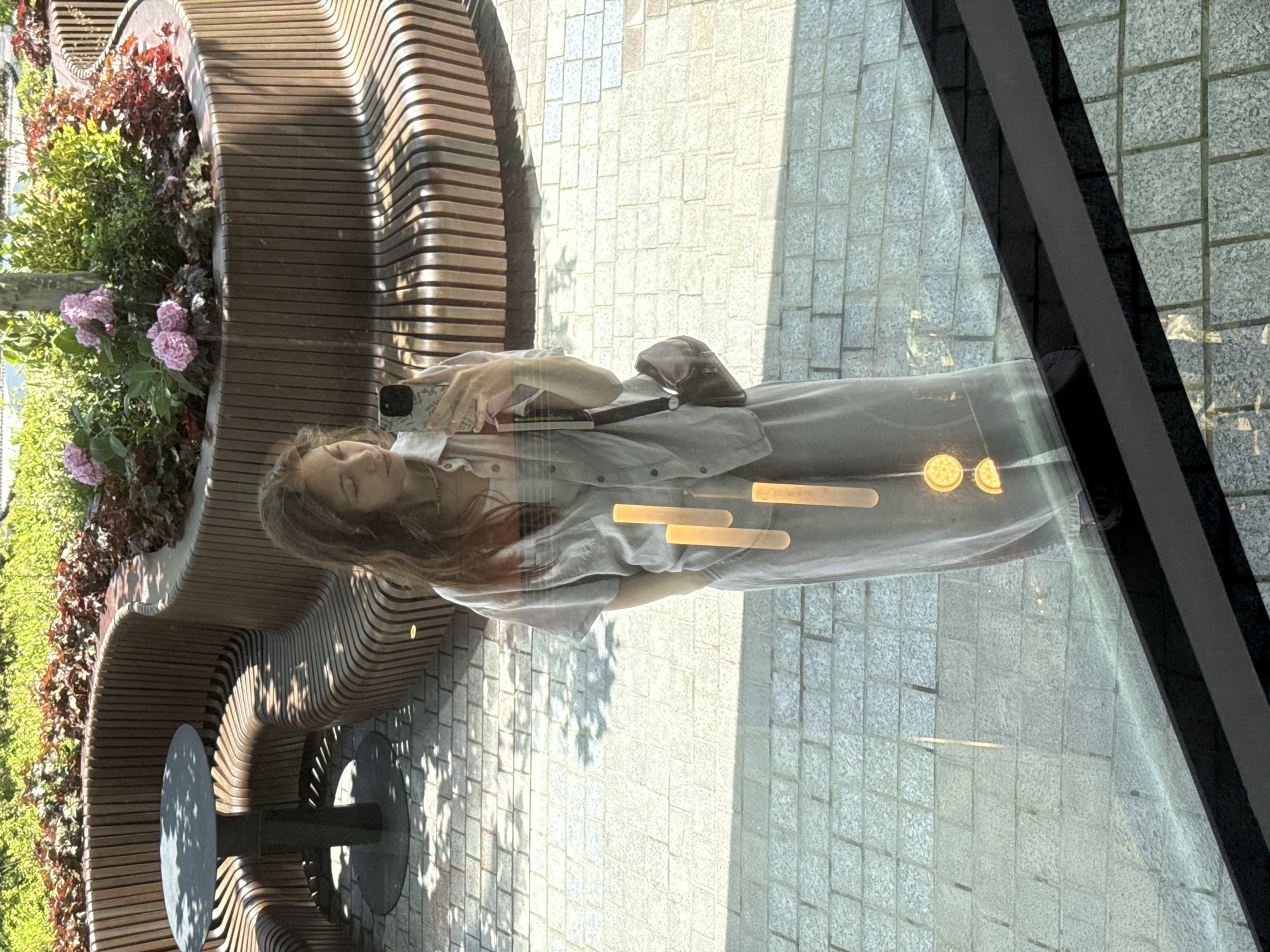 A woman is taking a selfie in a reflective surface, possibly a window or a polished floor, in an outdoor urban setting. She is wearing a light grey button-up shirt and matching wide-leg pants. A dark brown crossbody bag is slung over her left shoulder. The background shows a curved wooden bench with a black table in front of it, surrounded by greenery and pink flowers. The ground is paved with light-colored bricks. Sunlight casts shadows on the ground. The reflection also shows several vertically elongated, cylindrical light fixtures with glowing yellow elements.