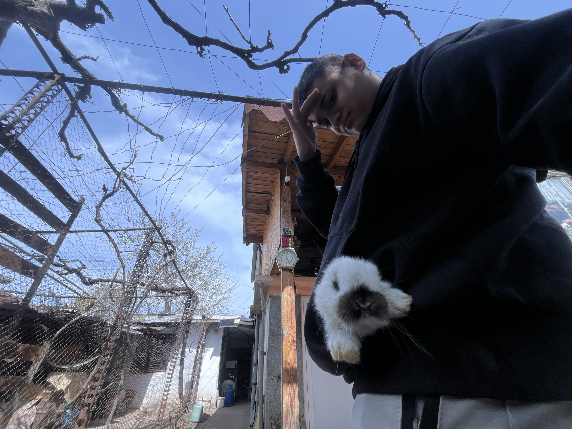A low-angle shot captures a young male outdoors in Lozarevo, Bulgaria. He wears a black hoodie and light-colored pants. He holds a small white rabbit with a dark face and ears in the front of his hoodie. The man has his other hand to his forehead. An old building with a red roof and a watch attached to the side is visible in the background. The sky is blue with scattered clouds. A dilapidated structure of rusted metal and chicken wire stands to the left, and bare branches overhang from the top of the frame. A partially visible alleyway to the left leads toward a storage room.