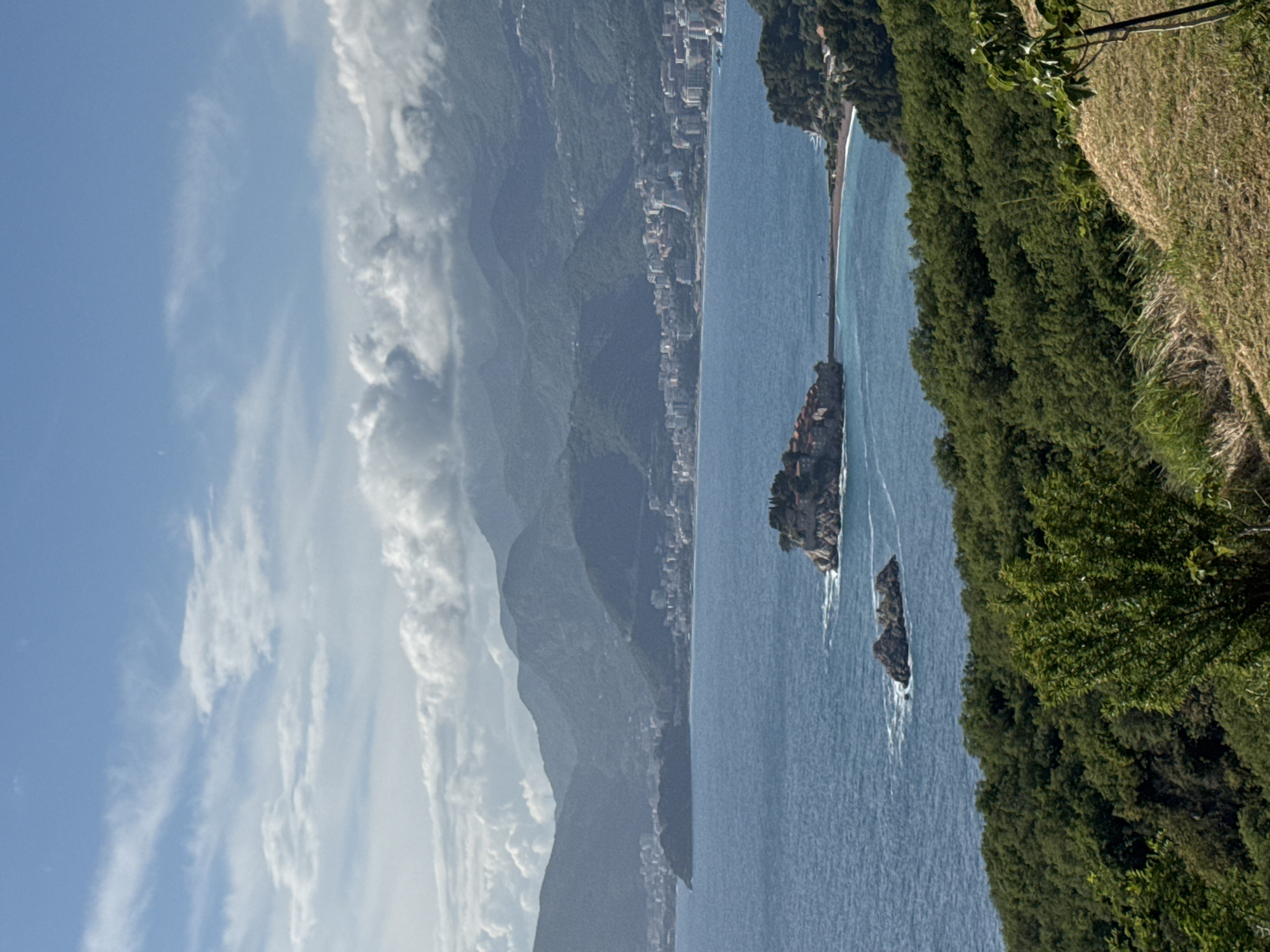 The image shows an elevated view of the Adriatic coast in Blizikuće, Montenegro. The perspective is oriented with the sky on the left and the shoreline on the right. Sveti Stefan, a small islet with buildings, is connected to the mainland by a narrow isthmus. Smaller rock formations are visible in the water near the islet.
The coastline is characterized by a stretch of buildings at the base of mountains. Lush green vegetation covers the foreground on the right. In the sky, there are streaks of wispy clouds against a blue background.