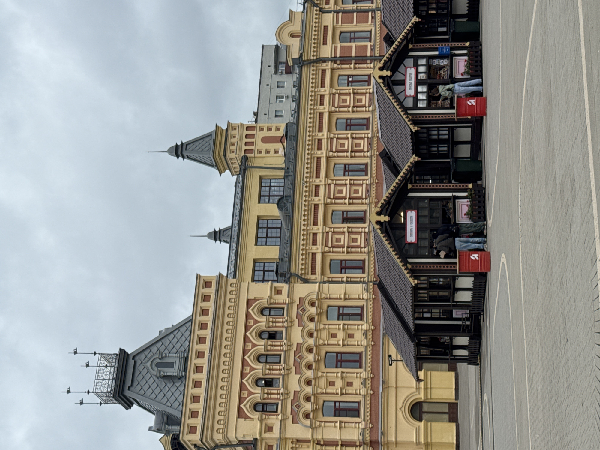 The image shows a view of the Nizhny Novgorod Kremlin in Nizhny Novgorod, Russia. In the foreground, there are several small market stalls with brown tiled roofs and dark wooden facades. Two individuals are standing at the stalls, interacting with the vendors. In front of one stall is a red cylindrical trash bin with a white "Я" symbol. The stalls are set up on a paved plaza. In the background, a large, ornate yellow and red building with multiple windows and architectural details stands prominently. The sky is overcast.