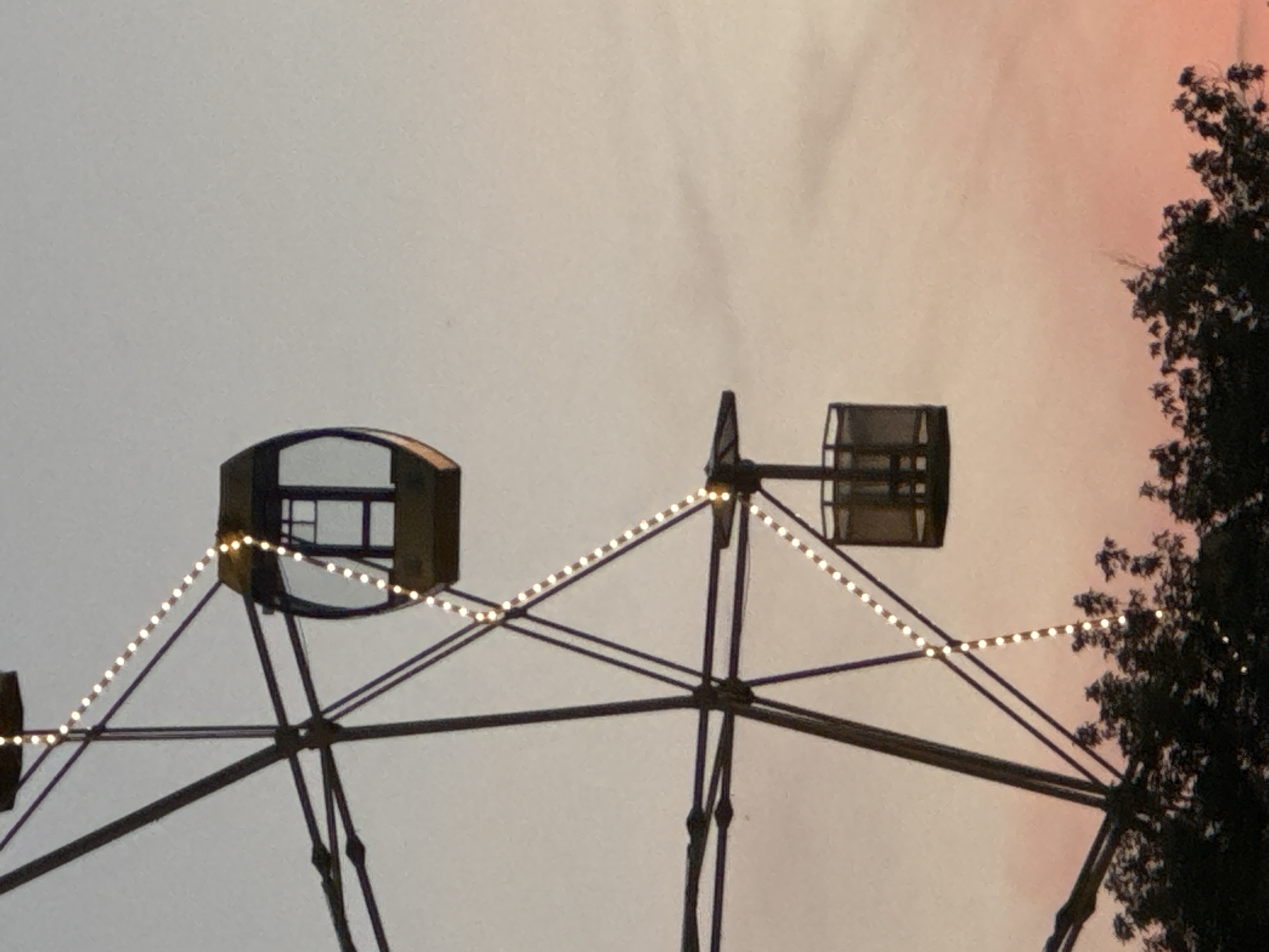 A Ferris wheel with a dark frame and passenger gondolas is silhouetted against a gradient sky transitioning from pale gray at the top to soft pink and orange at the bottom. String lights, resembling tiny white bulbs, are visible along the spokes of the Ferris wheel, creating a dotted line effect. Two passenger gondolas are visible; one is darker and boxier, while the other is lighter and more rounded with windows. The lower portion of the image shows the dark silhouettes of trees. The scene appears to be at dusk or dawn.
