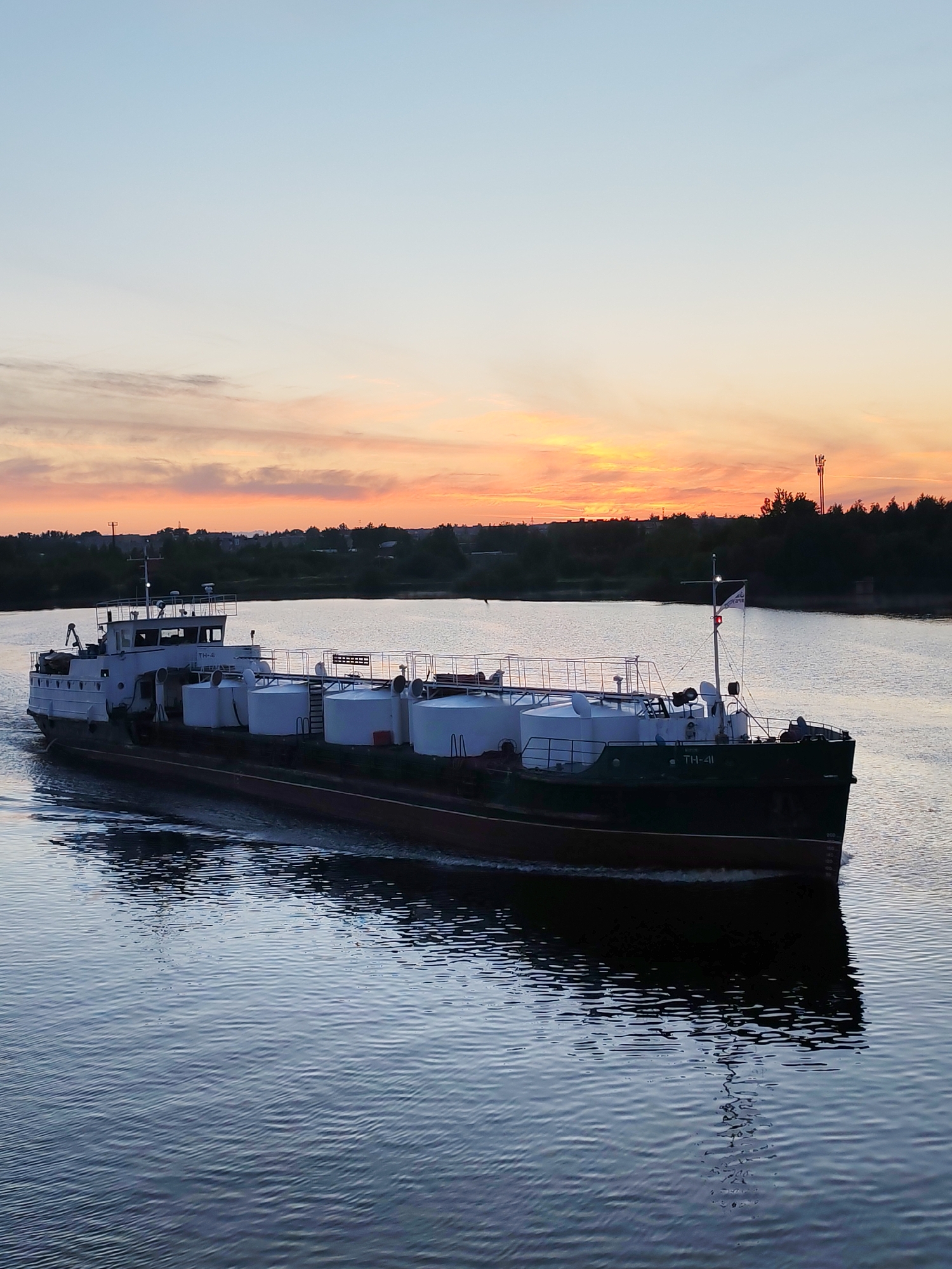 The image depicts a ship, possibly a tanker or cargo vessel, moving on a body of water at dusk or dawn. The ship is dark in color with a lighter-colored superstructure, and it appears to be carrying several large, white cylindrical tanks on its deck. A flag is visible on the ship's stern.
The background consists of a skyline with trees and some buildings silhouetted against a colorful sky with orange and pink hues. The water is dark and reflective, with visible ripples and waves indicating movement. There is a telecommunications tower visible on the right. The location appears to be a river or large canal in an area with vegetation along the banks. It's difficult to determine the specific location in the city of Unknown, Unknown without further information.