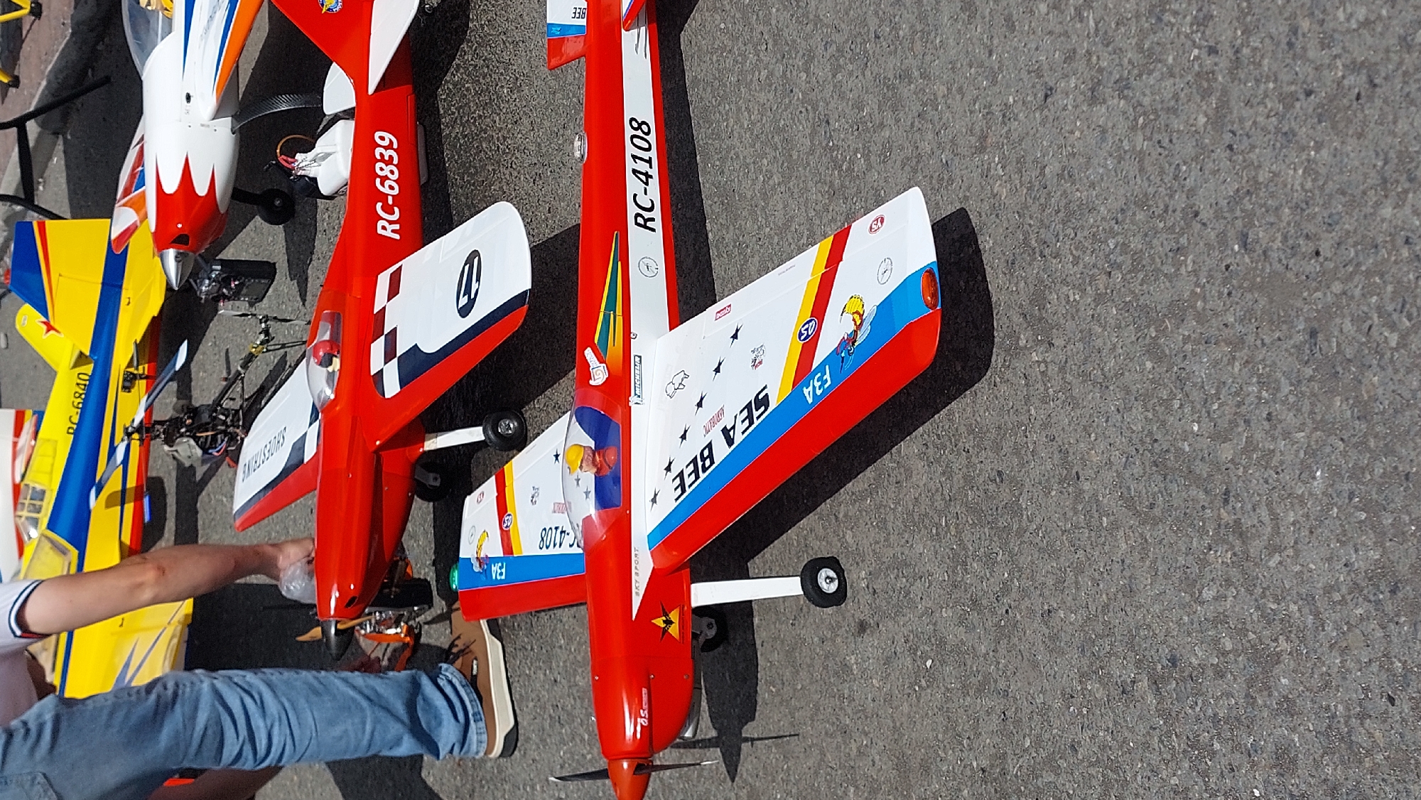 In Petropavlovsk-Kamchatsky, Russia, on an asphalt surface, a collection of model aircraft is displayed. In the foreground, a red and white model airplane, identified by "RC-4108" on its fuselage and "SEA BEE" on its wing, is positioned. Its propeller is visible, and a pilot figure is seated in the cockpit. Behind it, another red and white model aircraft, marked "RC-6839," is partially visible. Further back, a yellow and blue model airplane with the marking "RC-6840" is also on display. A small model helicopter is situated between the yellow and the red and white planes. A person's legs, clad in blue jeans and brown shoes, are visible on the left side of the image, suggesting they are interacting with or standing near the models. The scene appears to be an outdoor event or display, likely for hobbyists.