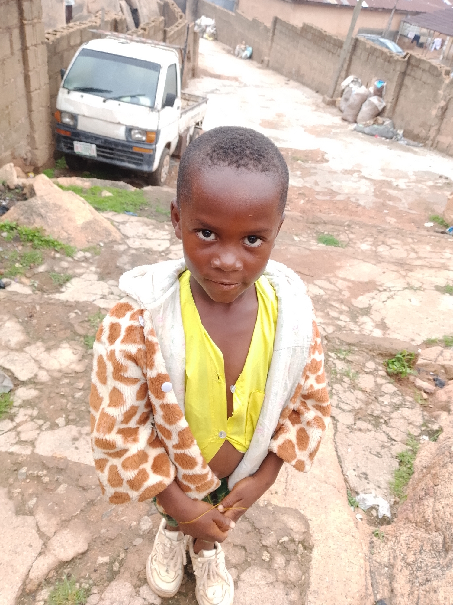 A young Black child stands outdoors, looking at the camera. The child is wearing a yellow shirt and a patterned jacket with brown and white spots. The child's hands are clasped together, and they are wearing white sneakers. The background shows a dirt path with scattered rocks and vegetation. In the distance, a white truck and a brick wall are visible, suggesting an urban or semi-urban environment. The lighting suggests it is daytime, possibly overcast.