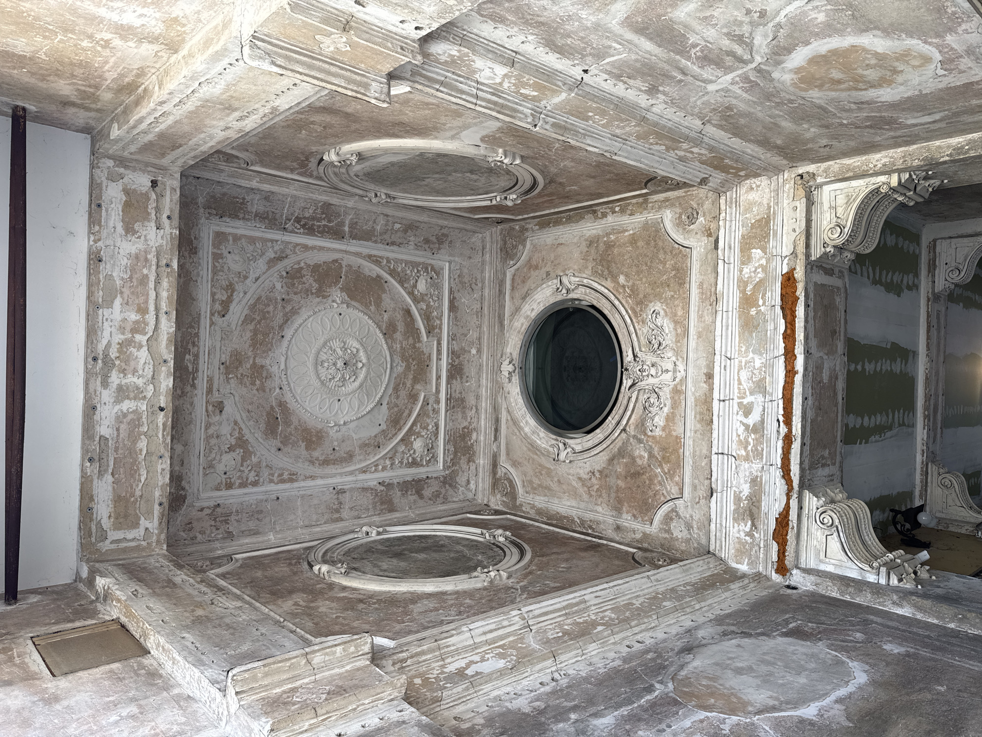 The image depicts an ornate, dilapidated room in Milan, Italy. The room features a heavily decorated ceiling with circular and square recessed panels, each adorned with intricate plasterwork and moldings. The walls are similarly decorated with plasterwork, but show significant signs of damage, including exposed brick and missing sections. The floor is marked with circular stains. An open doorway on the right side of the image reveals another room undergoing renovation, with bare walls and construction materials visible. No people are present in the scene.