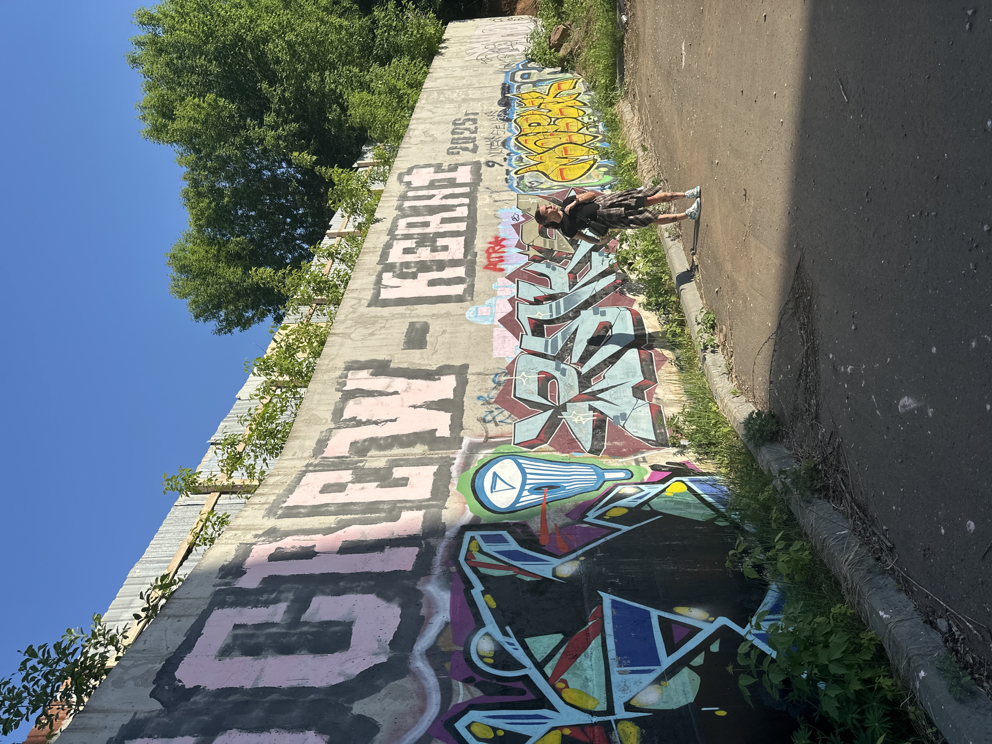 A medium-shot captures a male individual standing against a graffitied wall. He is wearing a dark t-shirt, plaid shorts, and light-colored sneakers. He has a backpack on and is angled slightly towards the viewer. The wall is concrete and covered in vibrant, multi-colored graffiti. Above the wall, there is foliage and trees, and a clear blue sky is visible. The surface where the individual stands is asphalt. The location is Izhevsk, Russia.