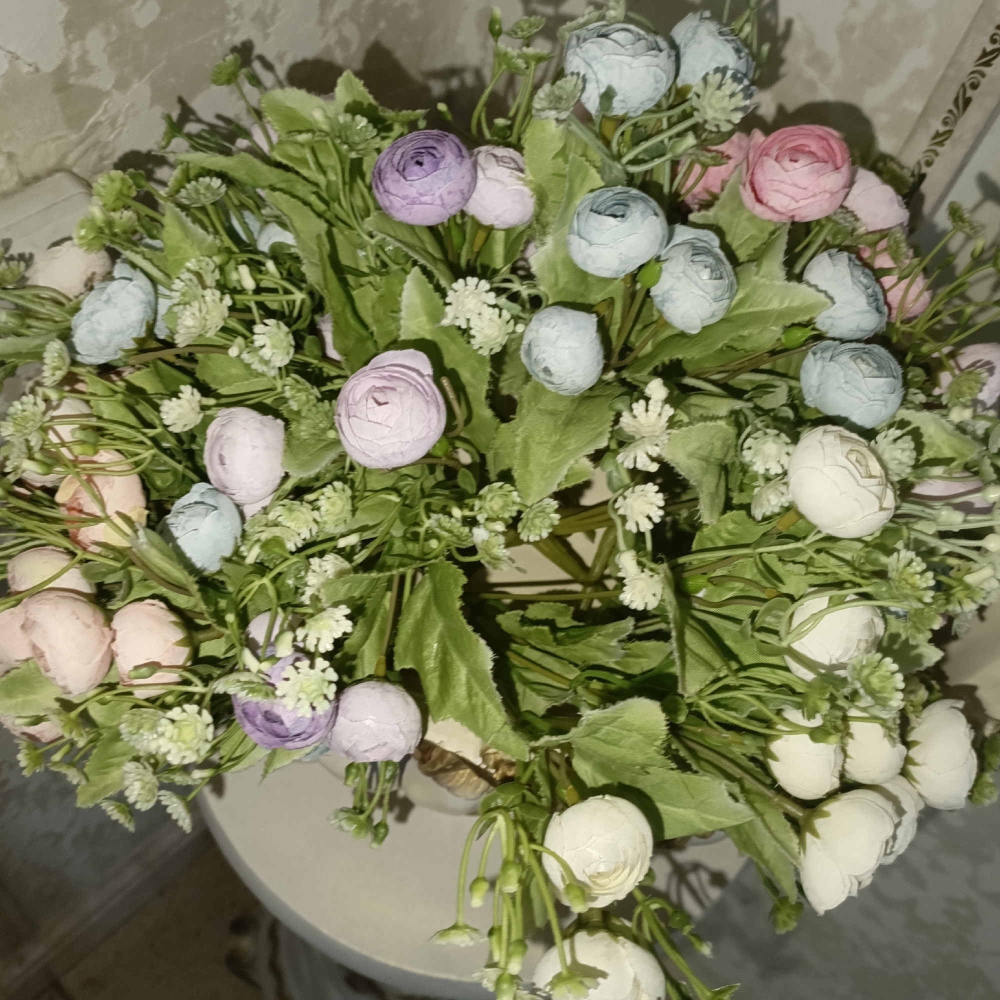 This is a close-up, top-down photograph of a floral arrangement composed of artificial flowers and greenery, likely situated indoors. The bouquet is dense and features a variety of flower shapes and sizes in pastel shades of pink, purple, blue, and white. Small, delicate white florets are interspersed throughout the arrangement. The backdrop appears to be a textured wall, possibly wallpapered, with a light beige or grayish hue. The flowers are arranged in a basket or pot, the top edge of which is visible at the bottom of the image, appearing to be made of a natural material like wicker. The lighting suggests an indoor setting, with shadows cast by the bouquet onto the surface it rests upon and the wall behind. No people are present in the image, and no specific location within Kenzhe, Russia, can be identified from the visual information provided.