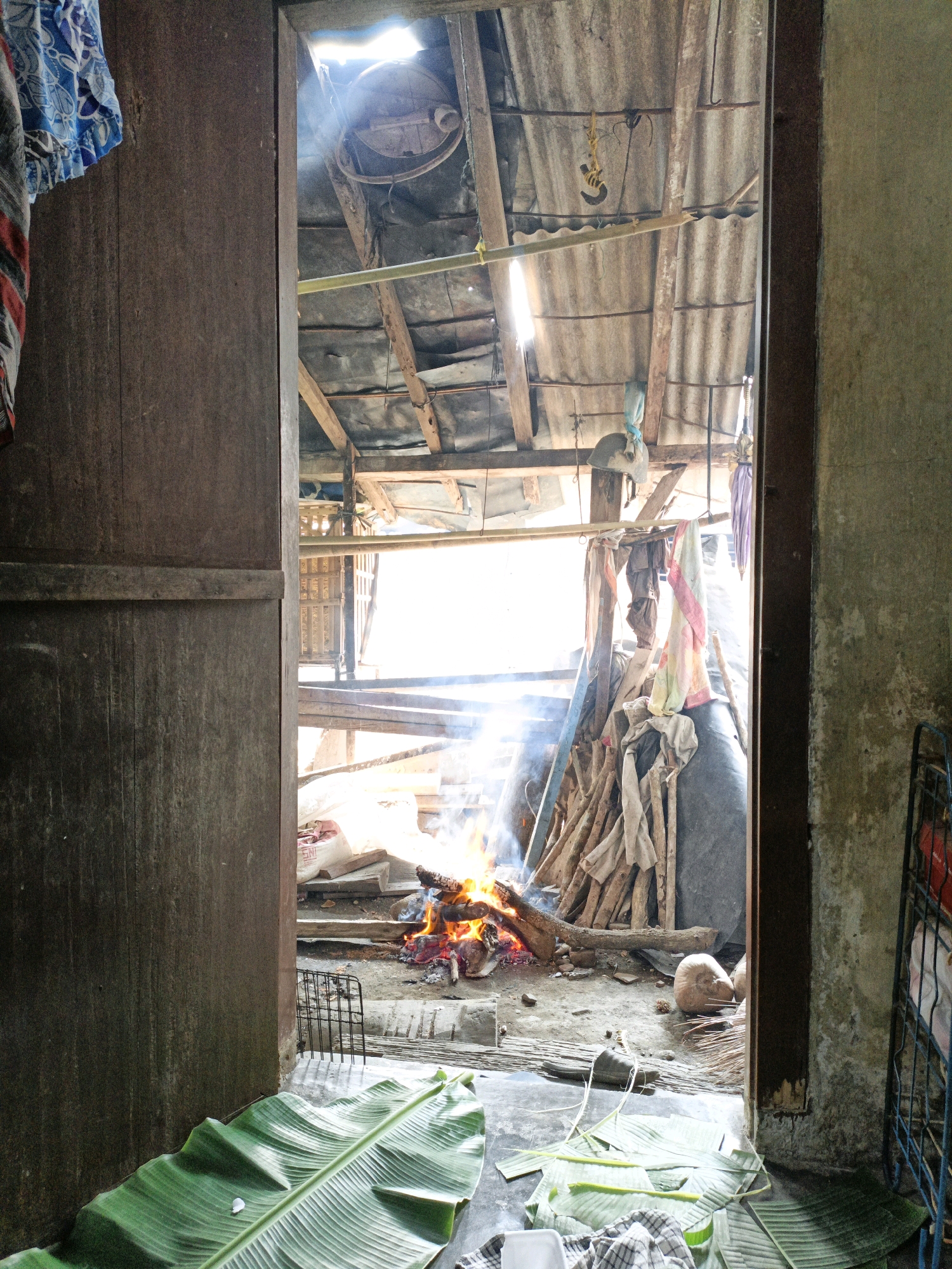 This is an eye-level, medium shot of a rustic, open-air structure, possibly a kitchen or workshop, in Bunut, Indonesia. The scene is viewed from a doorway, with a weathered wooden doorframe and a plank of wood on the left. Sunlight streams in from the open structure, illuminating a fire burning in the center of the dirt floor. Logs and branches are piled up to the right of the fire. Several pieces of fabric, including what appear to be towels and clothing, are draped over a wooden beam above the fire. Banana leaves are spread out on the floor in the foreground, some appearing to be freshly laid, while others are crumpled. There is a metal grate or cage to the left of the fire. In the background, there are more wooden structures and bundles of materials. The roof appears to be corrugated metal. The overall impression is one of a simple, functional living or working space, likely used for cooking or other domestic activities. There are no people visible in the image.
