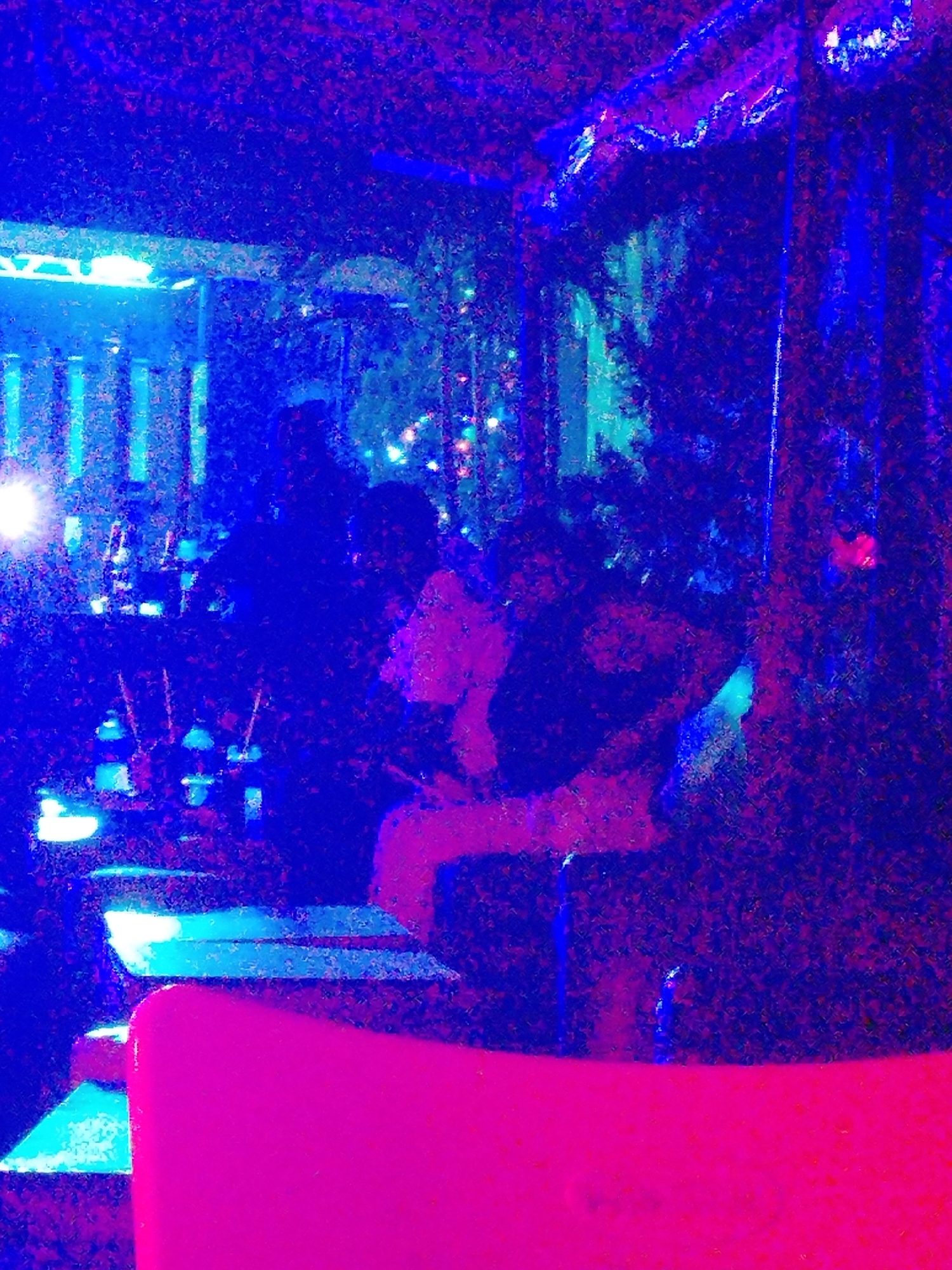 The image depicts an indoor space, likely a bar or restaurant, possibly in Yelwa, Nigeria, although this cannot be confirmed with certainty from the image alone. The lighting is primarily blue and magenta, creating an artificial ambiance. Several people are present, though their features are obscured by the low light and image quality. Some individuals appear to be seated at tables. There are bar-like fixtures and possibly decorative elements such as plants visible in the background. In the foreground is what seems to be a pink chair.