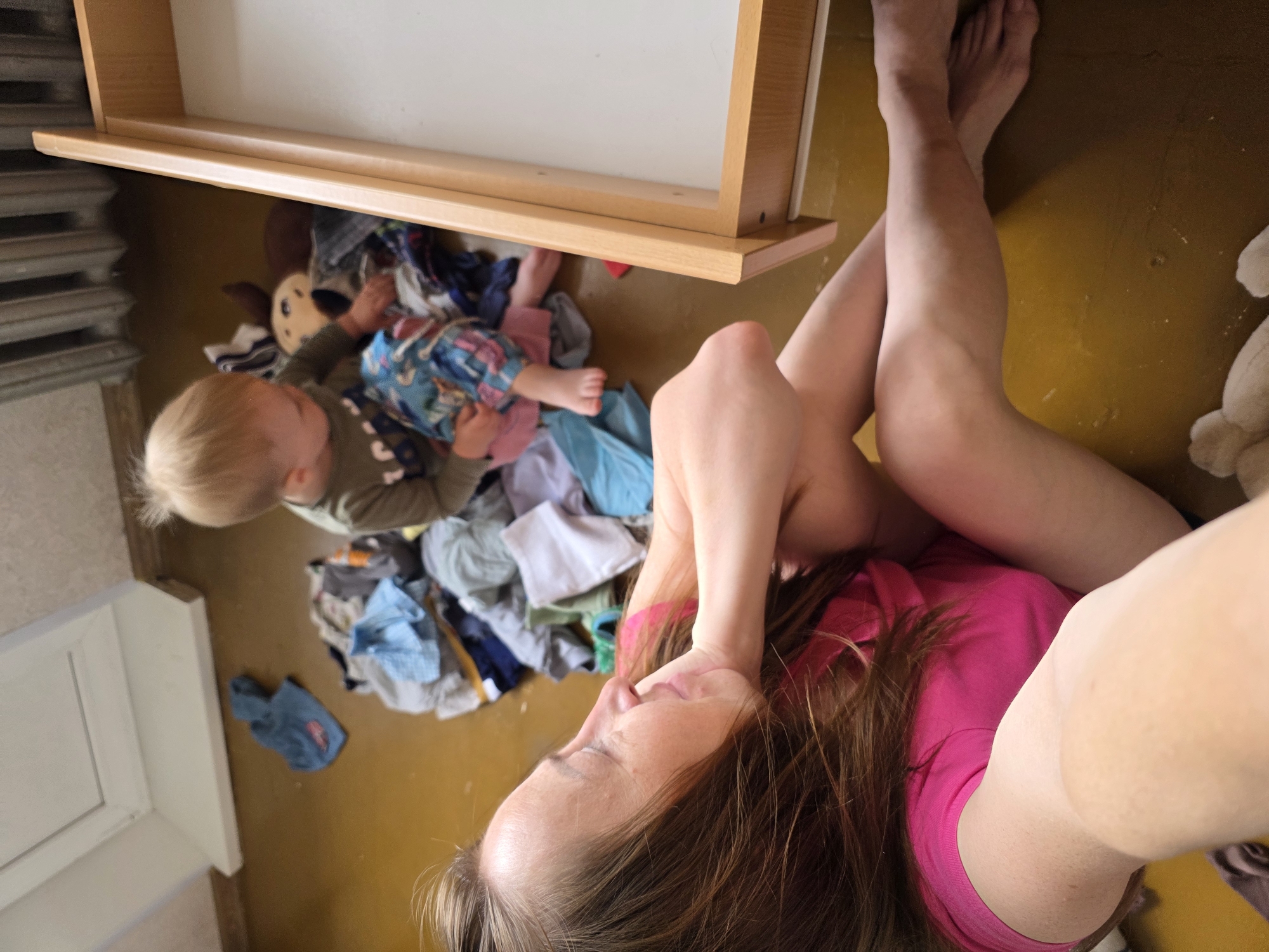 An adult female and a young child are depicted indoors on a yellowish-brown floor, which is identified as being in the city of Chaikovsky, Russia. The woman, with long brown hair and wearing a bright pink t-shirt, is reclined on the floor, supporting her head with her right hand and looking to her right with a slight smile. Her bare legs and feet are visible. She appears to be taking a photograph with her left arm extended towards the camera. A blonde-haired toddler, wearing a dark olive green long-sleeved shirt and patterned blue and pink shorts, is seated on the floor amidst a substantial pile of assorted clothing items. The child is looking down and appears to be interacting with the clothes. Behind the child, a brown and beige stuffed animal is partially visible within the clothing pile. To the right of the child, a light-colored wooden drawer is open and empty, revealing a white interior. In the upper background, behind the child, a metallic radiator with vertical fins is visible. The upper left portion of the image shows a white baseboard or trim where a wall meets the floor.