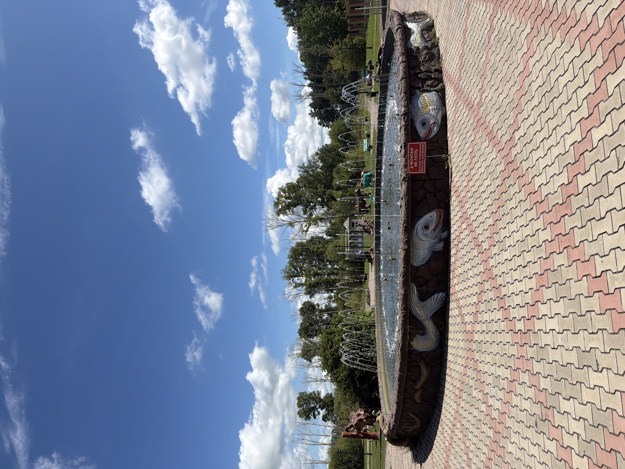 The image shows a circular fountain with water jets in a park setting, under a bright blue sky with scattered white clouds. The fountain's rim is decorated with sculpted fish. A red sign on the rim reads "В ФОНТАН НЕ ЛЕЗЬ! ВЫСОКОЕ НАПРЯЖЕНИЕ! ОПАСНО ДЛЯ ЖИЗНИ!", which translates to "Do not climb into the fountain! High voltage! Dangerous to life!". In the background, there are trees, a playground area with a slide and figures, and a few people visible. The ground around the fountain is paved with interlocking bricks in a pattern of light and dark red and beige. The overall scene depicts a public outdoor recreational area.