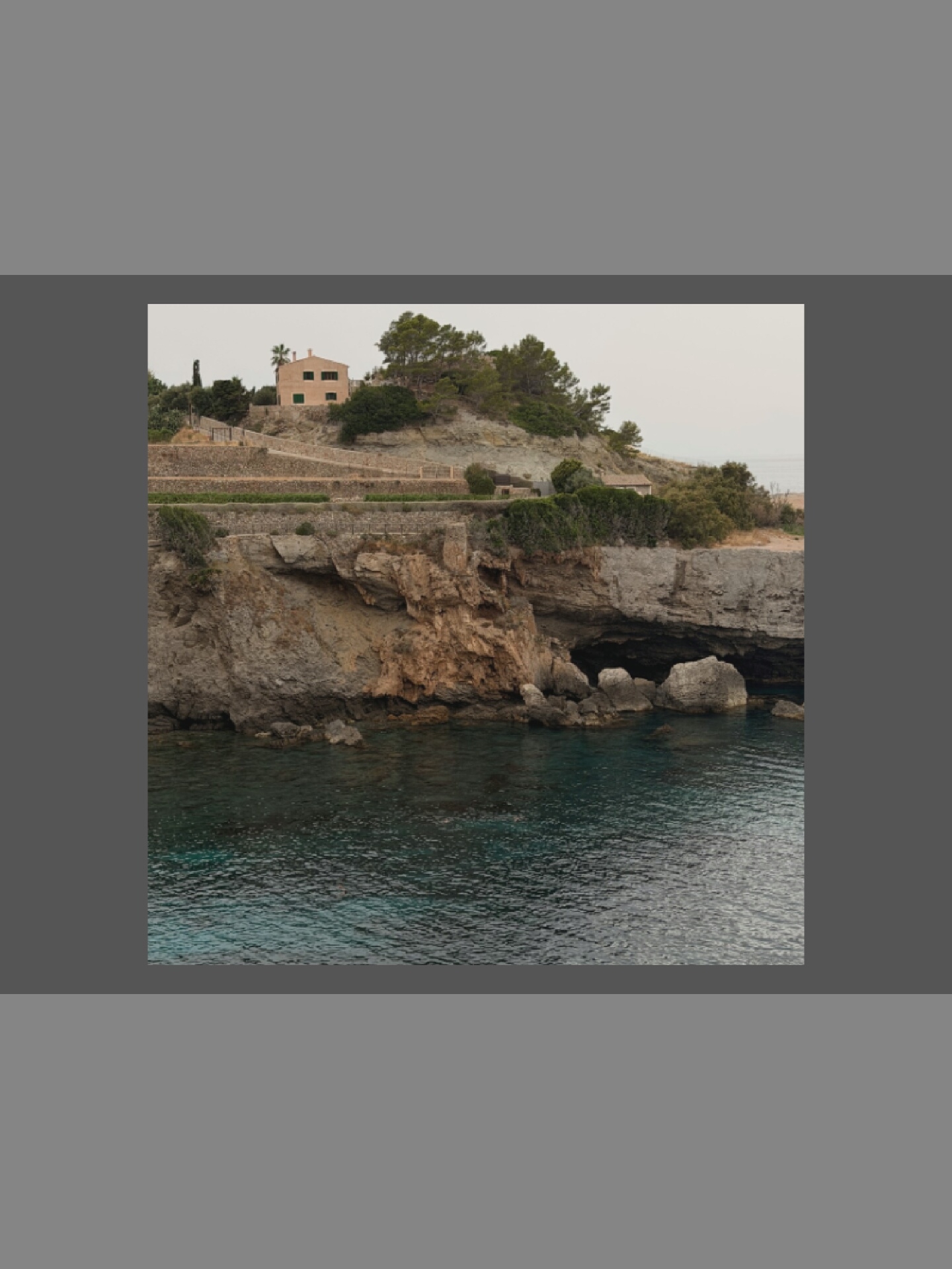 A wide shot captures a rugged, rocky coastline with a small, light-colored house perched on a hill overlooking the water. The house has a terracotta roof and green shutters. Lush green trees and shrubs surround the house, clinging to the rocky terrain. A stone wall and pathway lead up to the property. Below the cliff, clear turquoise water laps against the rocks, revealing submerged boulders. The sky is overcast with a soft, diffused light.