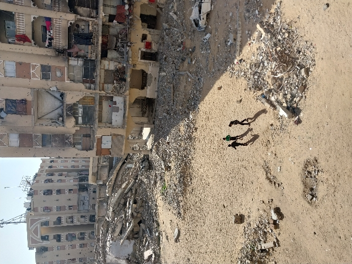 The image depicts a devastated urban landscape in Khan Yunis, Palestinian Territory, under a clear sky. In the foreground, three individuals, likely children, walk from right to left across an expanse of light brown sand. They cast distinct shadows to their left. The sandy ground is uneven, with a small depression visible in the lower-left and scattered piles of concrete rubble and debris throughout.
In the midground, the scene is dominated by severely damaged multi-story apartment buildings. The building on the right, closest to the foreground, shows extensive structural compromise across its visible six to seven floors. Its facade is pockmarked with numerous impact points, and most windows are either shattered or entirely missing, revealing dark, damaged interiors. Some balconies remain, with red and dark laundry items hanging from one on an upper floor. The lower levels of this building are particularly heavily damaged, with large sections of walls missing and foundations exposed, creating open spaces filled with rubble. To the right of the walking individuals, a heavily damaged white vehicle is partially buried in a large pile of debris.
Further in the background, another large, multi-story building displays similar damage, including numerous facade impacts. This building features distinctive arched architectural elements near its roofline, topped by communication antennas. Between these primary buildings, the ground is completely obscured by vast piles of collapsed concrete, twisted rebar, and other construction materials, indicating widespread destruction of intermediary structures.