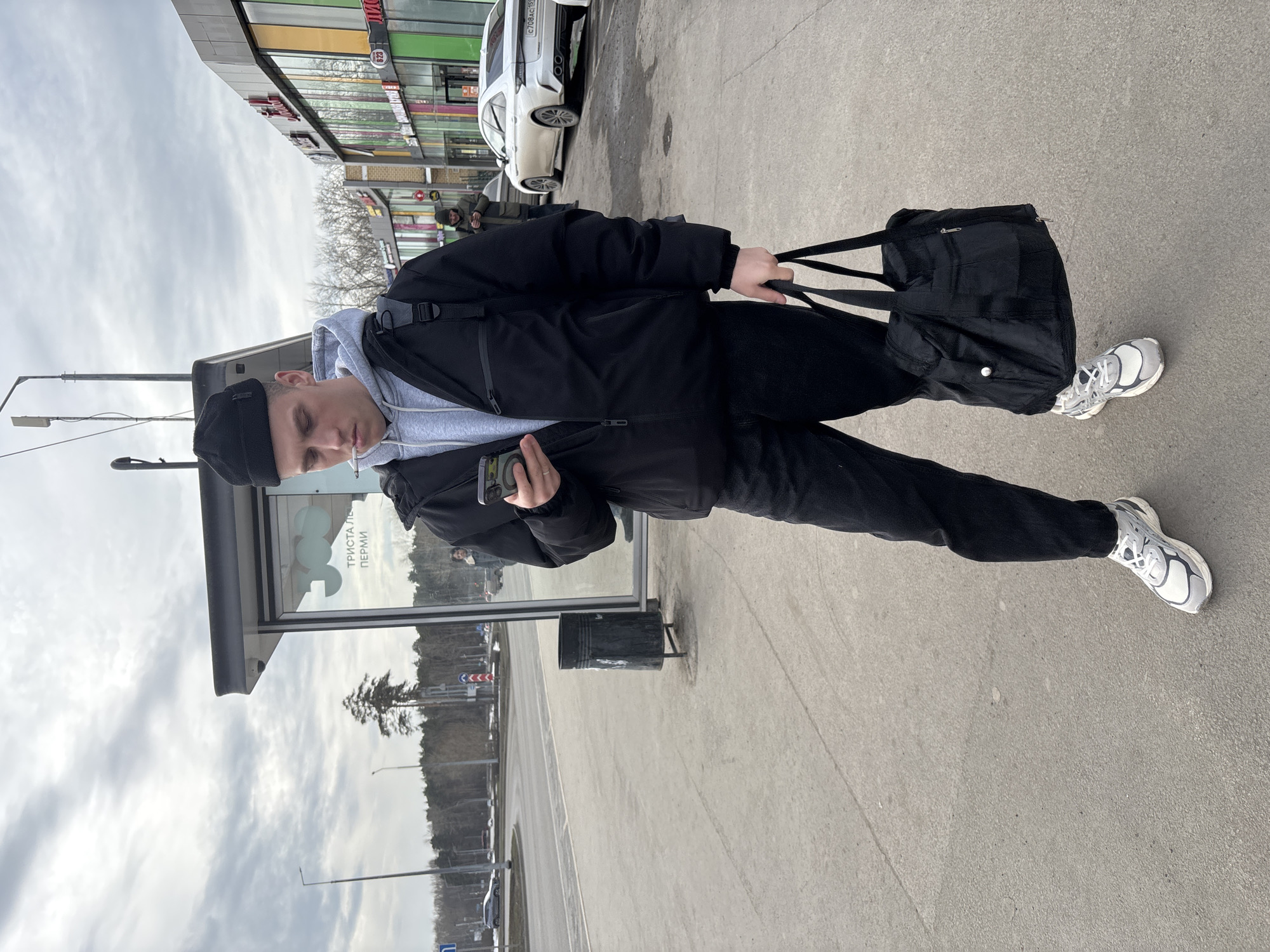 A young man stands at a bus stop in Perm, Russia, holding a black bag in his right hand and a cell phone in his left. He wears a black beanie, black jacket, grey hooded sweatshirt, black pants, and white sneakers. A cigarette is in his mouth. The bus stop has glass walls with writing in Russian visible on them. A black metal trash can stands nearby. A street and trees are visible through the bus stop's glass. In the background, across the street, is a shopping center and a white car parked.