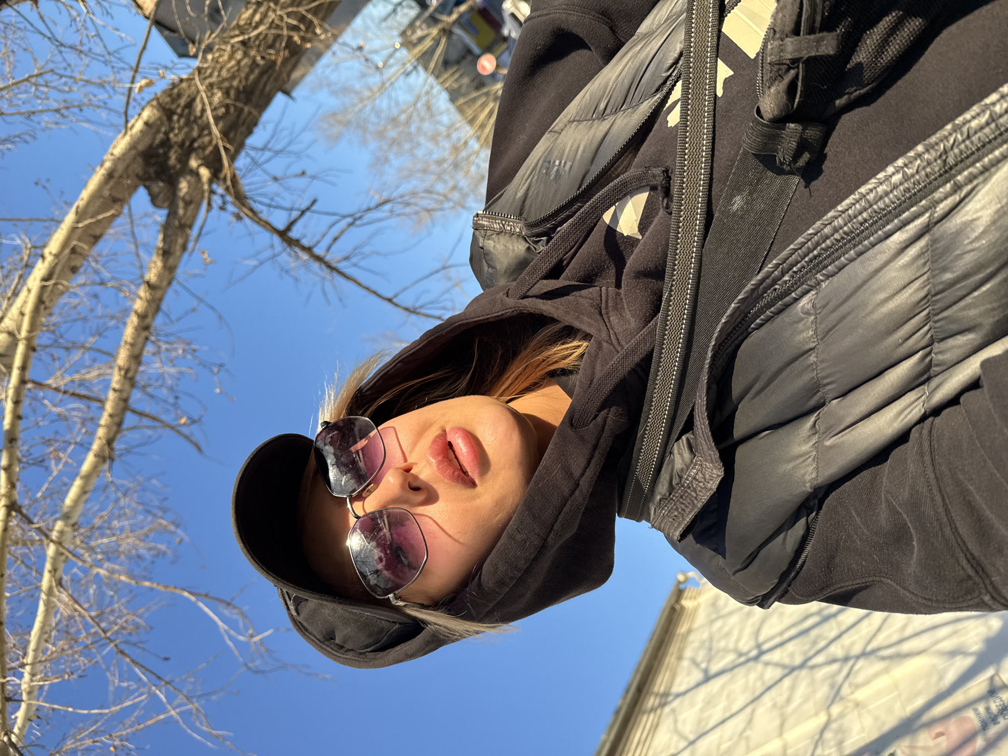 A woman wearing sunglasses and a black hooded jacket stands outside under a clear blue sky. A bare tree with light-colored bark is visible in the background, along with a building or structure in the distance. Based on the bare tree, it is likely late fall, winter or early spring. Without further information, pinpointing the exact location in Borzya, Russia, is not possible.