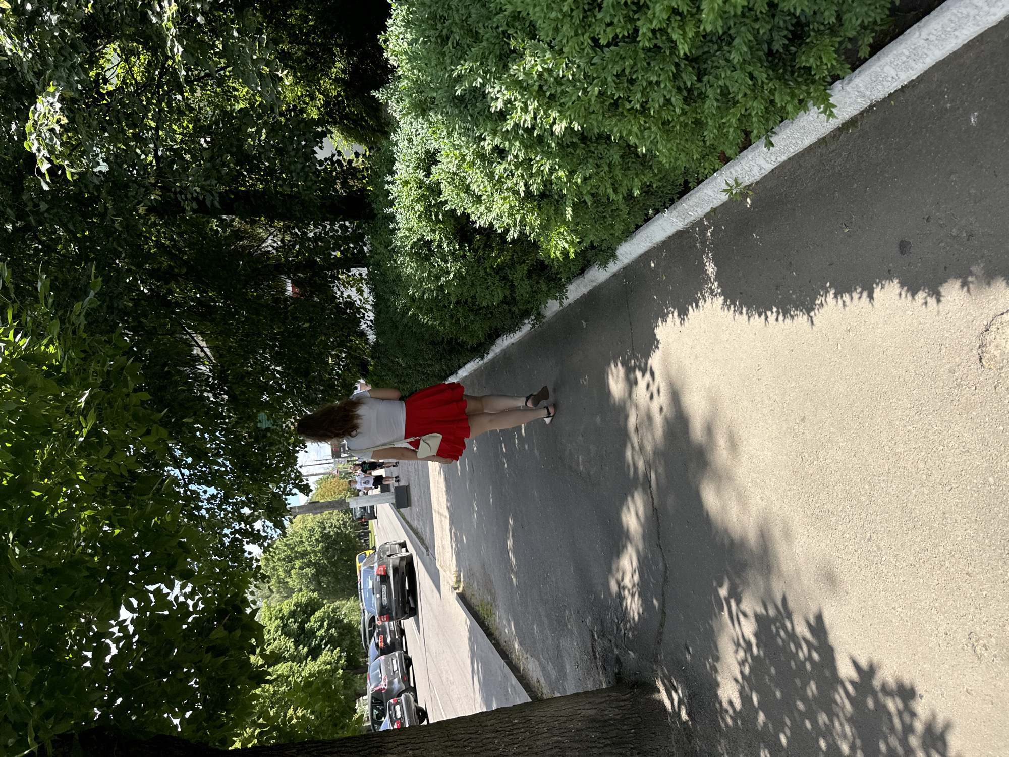 A woman with long brown hair walks away from the camera down a concrete sidewalk. She is wearing a white t-shirt and a red skirt. She has a small white purse slung over her shoulder. She is walking on the right side of a street in Sarny, Ukraine. The street is lined with green bushes and trees, with cars parked and driving on the road. Shadows from the trees are cast on the sidewalk.