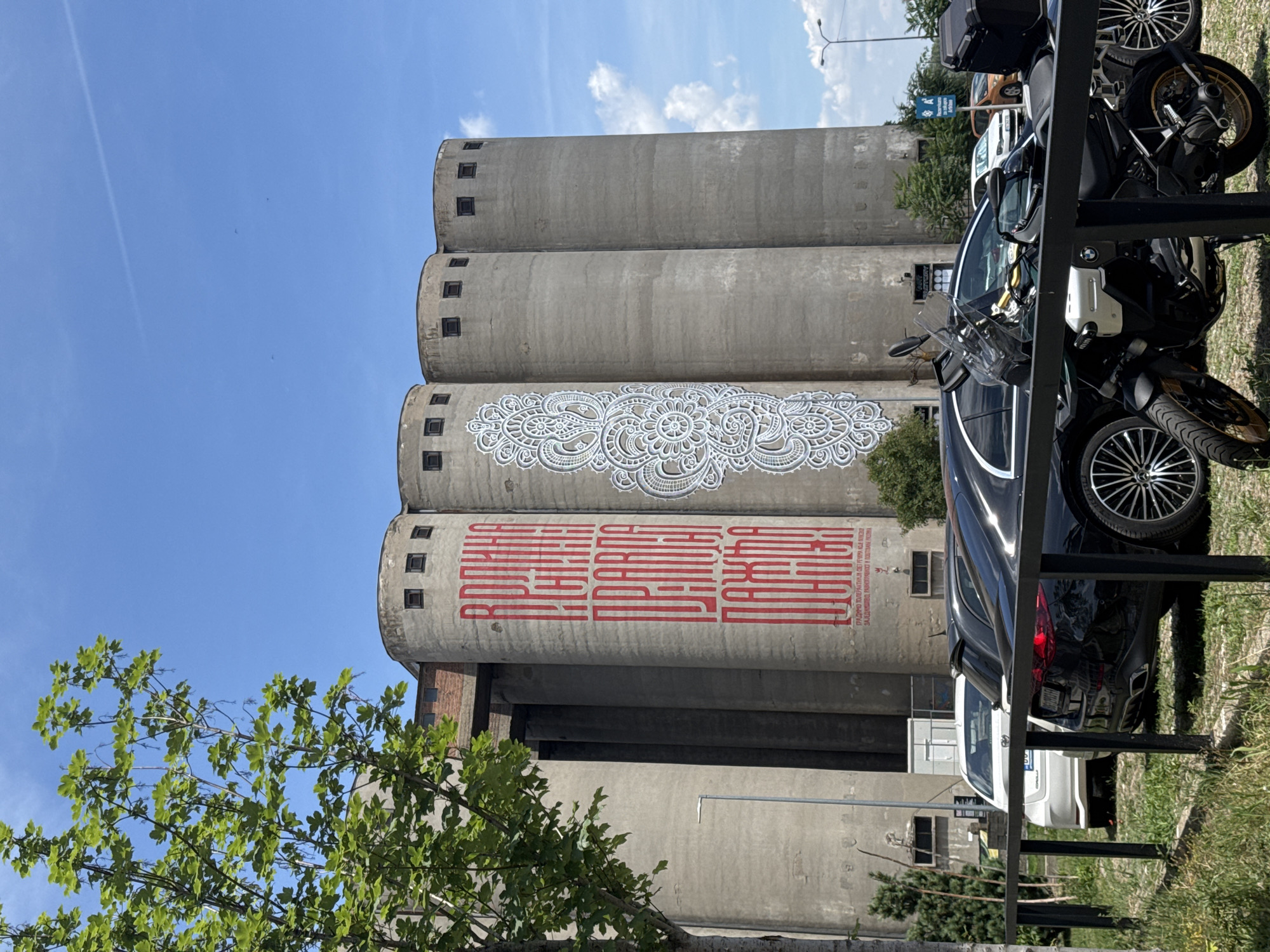 A multi-story industrial building, likely a former silo, dominates the frame, its cylindrical concrete structures adorned with large-scale artwork. The leftmost silo features vertical red lettering, while an adjacent silo displays an intricate white lace-like pattern. The building is situated in a metropolitan area, evidenced by parked cars and a motorcycle in the foreground. The environment suggests an urban setting, possibly Belgrade, Serbia, given the presence of Cyrillic script on the building, though this cannot be definitively confirmed without further context. The sky is a clear blue with scattered clouds, indicating fair weather. The foreground is occupied by a black car and a motorcycle parked on a patch of grass behind a black metal fence. A white car is also visible behind the black car. Trees and streetlights are present, suggesting an outdoor public space.