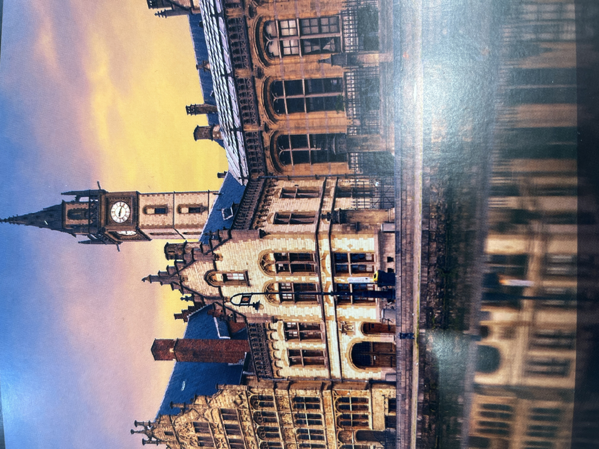 The image displays a picturesque canal in front of historic European architecture, possibly in a city like Ghent, Belgium, not Kharkiv, Ukraine. The buildings are made of light-colored stone and feature intricate details, including ornate windows and tiered rooftops. A tall clock tower with a visible clock face stands prominently to the right of center. The sky transitions from a deep blue at the top to a warm orange and yellow, suggesting either sunrise or sunset. The water in the canal reflects the buildings and the sky, creating a symmetrical and serene scene. There are no people or overt signs of activity visible.