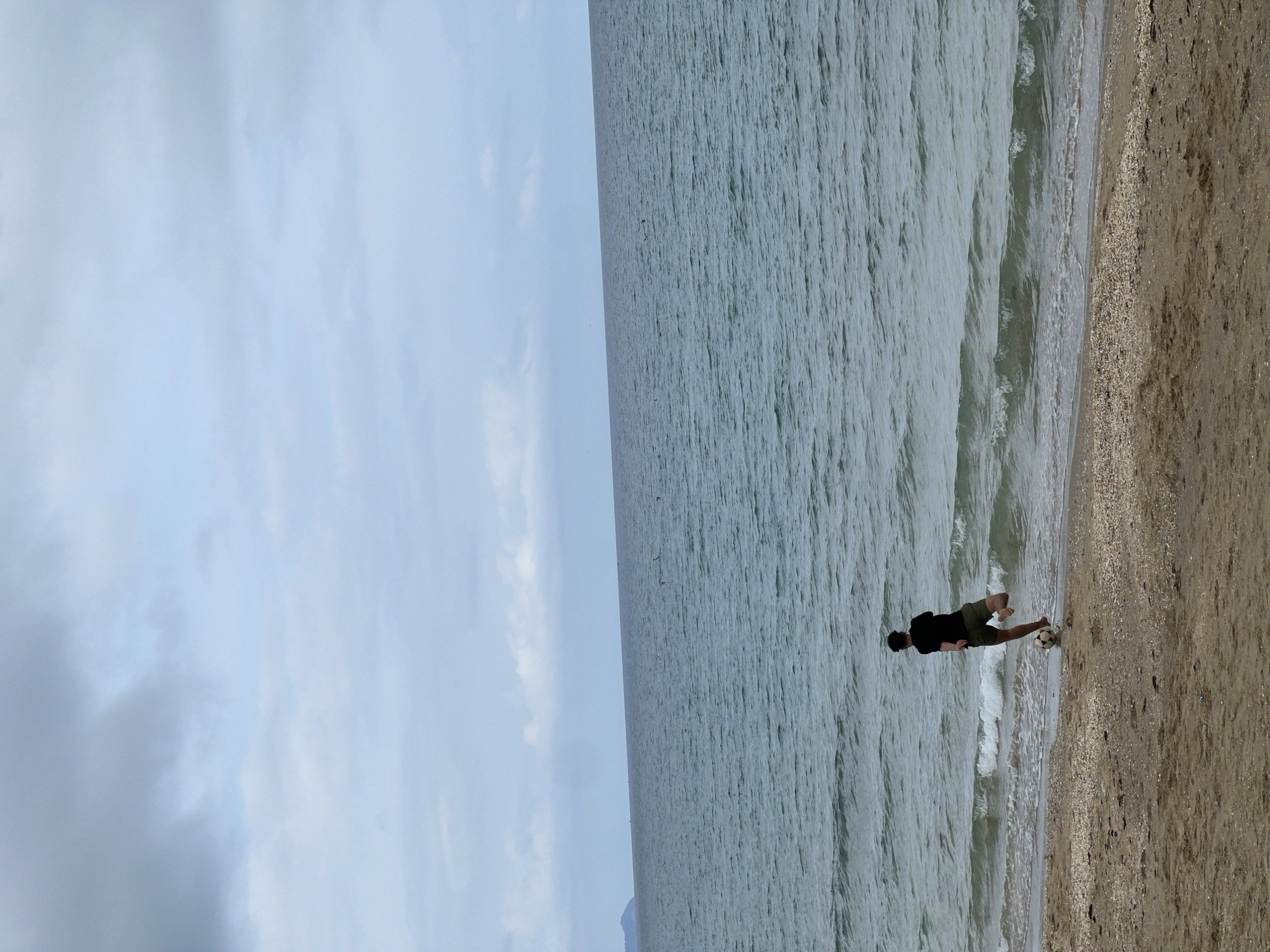 The image shows a coastal scene in M'Diq, Morocco. A person wearing a dark shirt and shorts is playing with a soccer ball on the beach. The beach consists of light-colored sand and small shells. The water is a muted green-grey, with small waves breaking on the shore. The sky is overcast with light grey clouds.