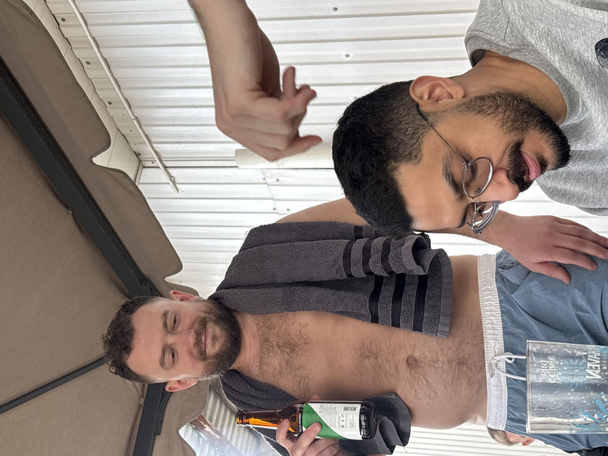 Two men are visible in the frame. One man, partially nude except for light blue shorts and a grey striped towel draped around his neck and shoulders, is holding a dark bottle. A second man, wearing a grey t-shirt and round glasses, appears to be seated or standing beside him. An additional arm is visible between the two men. A glass of liquid and part of an umbrella are also visible. The setting appears to be outdoors. There is no information in the image to indicate that it was taken in Krasnoyarsk, Russia.