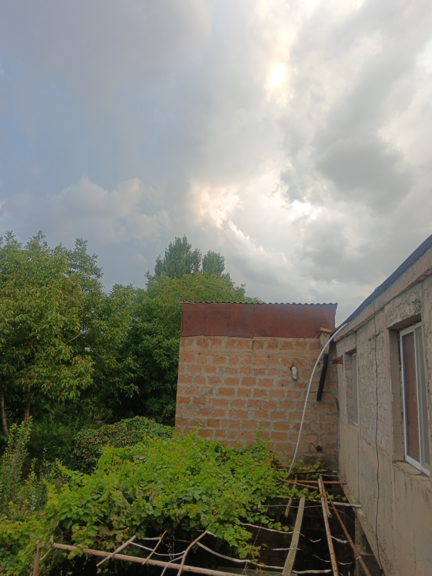 The image captures an outdoor scene with a cloudy sky featuring a sunbeam breaking through. In the foreground, there is a section of a building with a concrete wall and windows, adjacent to a brick structure topped with a corrugated metal roof. Lush green foliage, including trees and vines, occupies the left and lower portions of the frame. The overall setting appears to be a rural or semi-urban environment, consistent with a location in Aragatsavan, Armenia. There are no people visible in the image. The scene depicts a static outdoor environment with architectural elements and natural vegetation under an overcast sky.